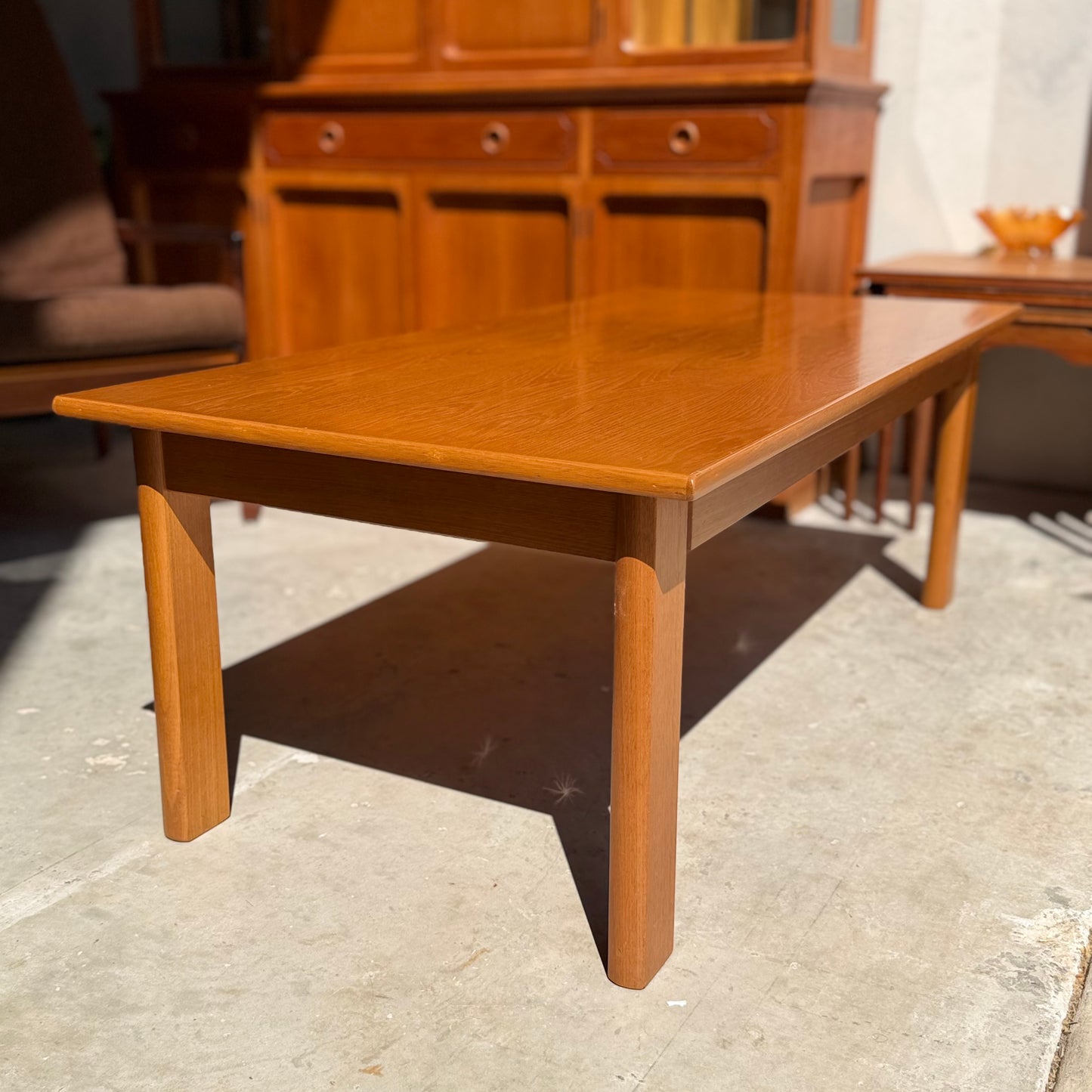 A Mid Century Teak Danish Style Coffee Table – Circa 1970s (Revive) with rounded legs sits on a concrete floor in sunlight, with a wooden cabinet and Scandinavian vintage furniture in the background.