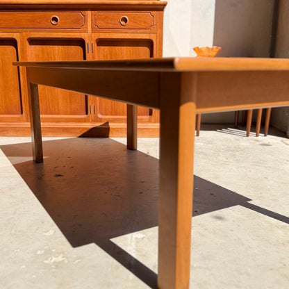 A Mid Century Teak Danish Style Coffee Table – Circa 1970s (Revive) rests on a sunlit concrete floor, casting a defined shadow, while behind it stands a matching sideboard topped with an orange decorative bowl.