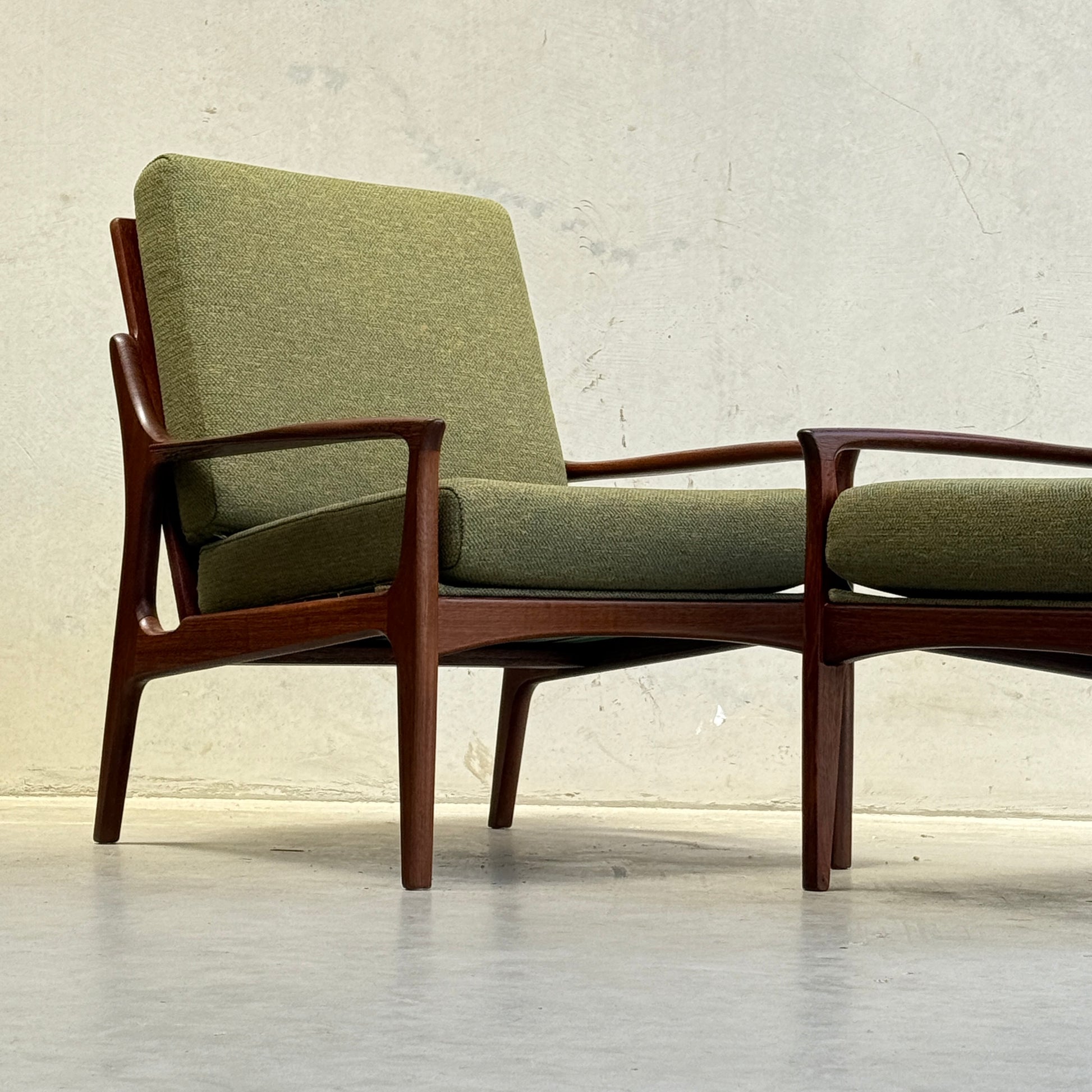 The Don Rex Armchair - Green Forest (Rediscover) features a minimalist mid-century modern design with angled legs, wooden frame, and green cushions, inspired by Danish modernism. It sits against a plain, light-colored wall and floor.