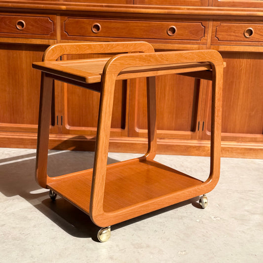 The Mid-Century Noblett Drinks Trolley – Removable Tray & Original Castors (Revive) features rounded edges and two teak shelves, standing on a sunlit concrete floor in front of matching wood cabinetry.