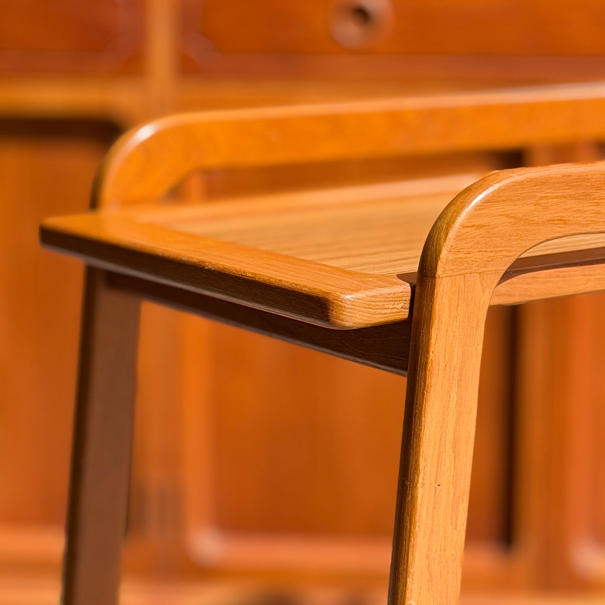 Close-up of a wooden chair with armrests and smooth edges, finished naturally. In the softly blurred background is a Mid-Century Noblett Drinks Trolley – Removable Tray & Original Castors (Revive), echoing the chair’s elegant style.