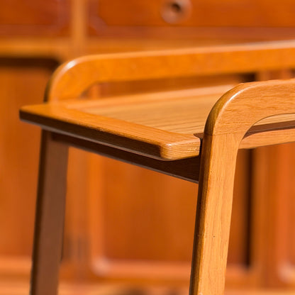 Close-up of a wooden chair with armrests and smooth edges, finished naturally. In the softly blurred background is a Mid-Century Noblett Drinks Trolley – Removable Tray & Original Castors (Revive), echoing the chair’s elegant style.