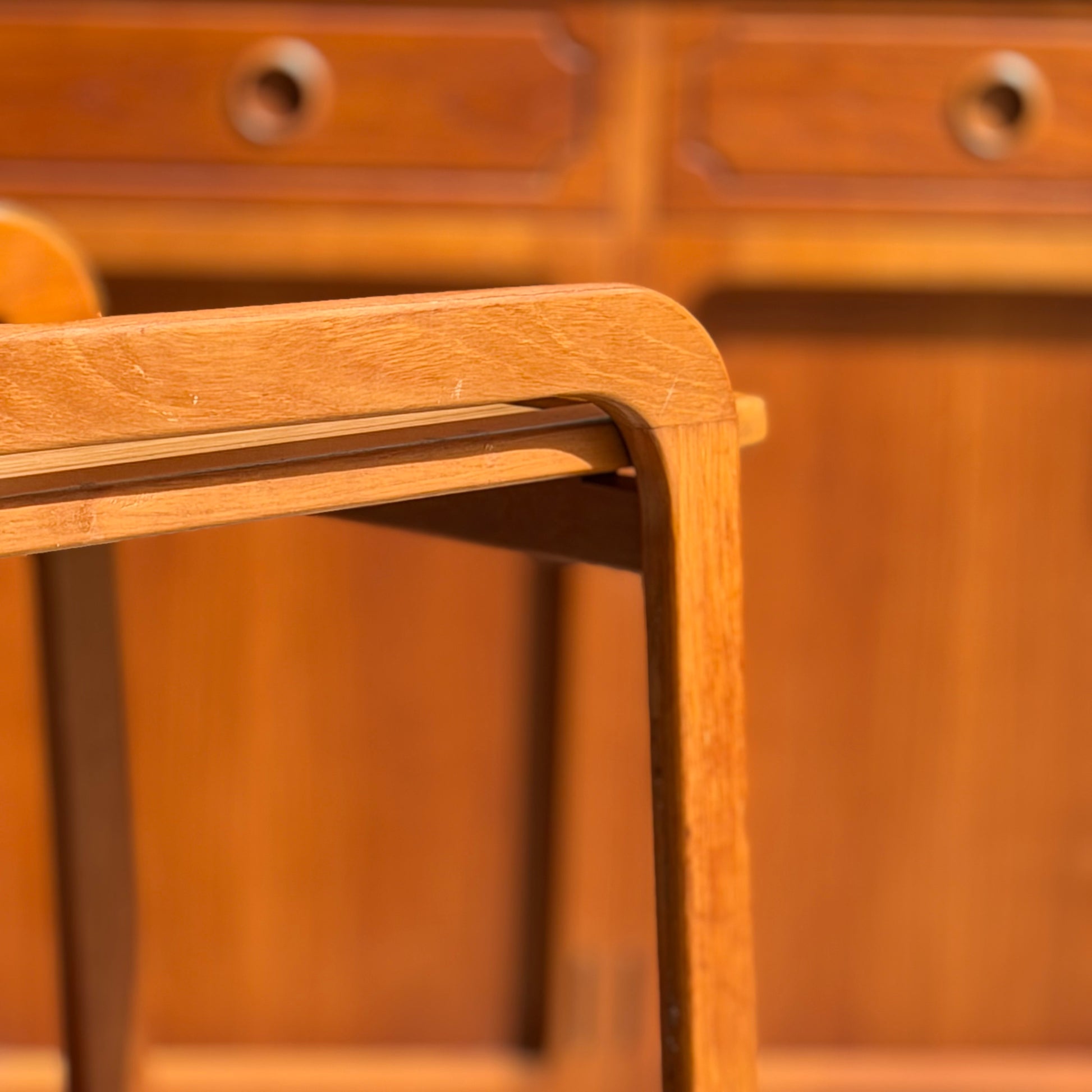 A close-up of a wooden chair leg and armrest, with a blurred Mid-Century Noblett Drinks Trolley – Removable Tray & Original Castors (Revive) featuring round handles in the background. Warm lighting highlights the wood grain and texture.