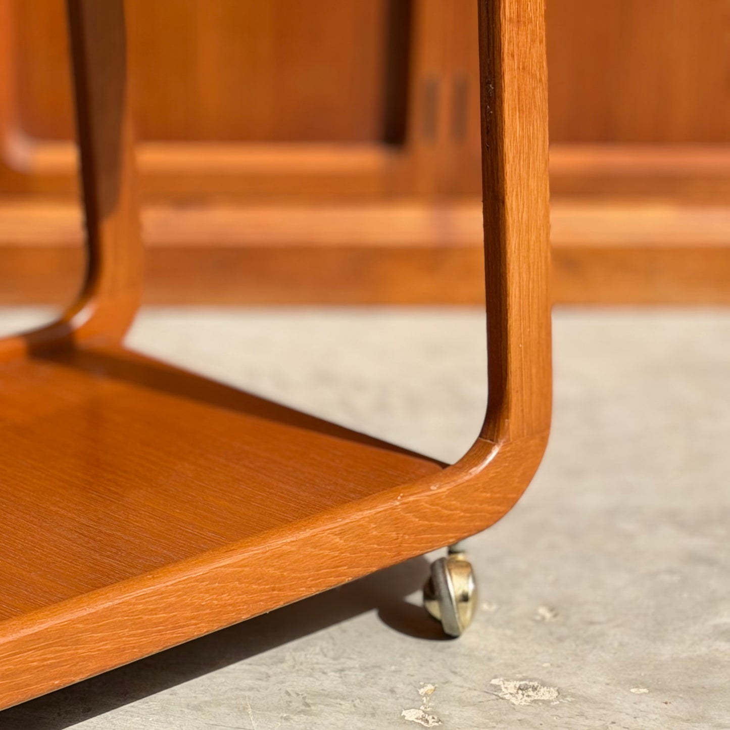 Close-up of the Mid-Century Noblett Drinks Trolley – Removable Tray & Original Castors (Revive), showing its rounded edges and castor, sunlit wood grain on a concrete floor. A blurred wooden cabinet appears in the background.