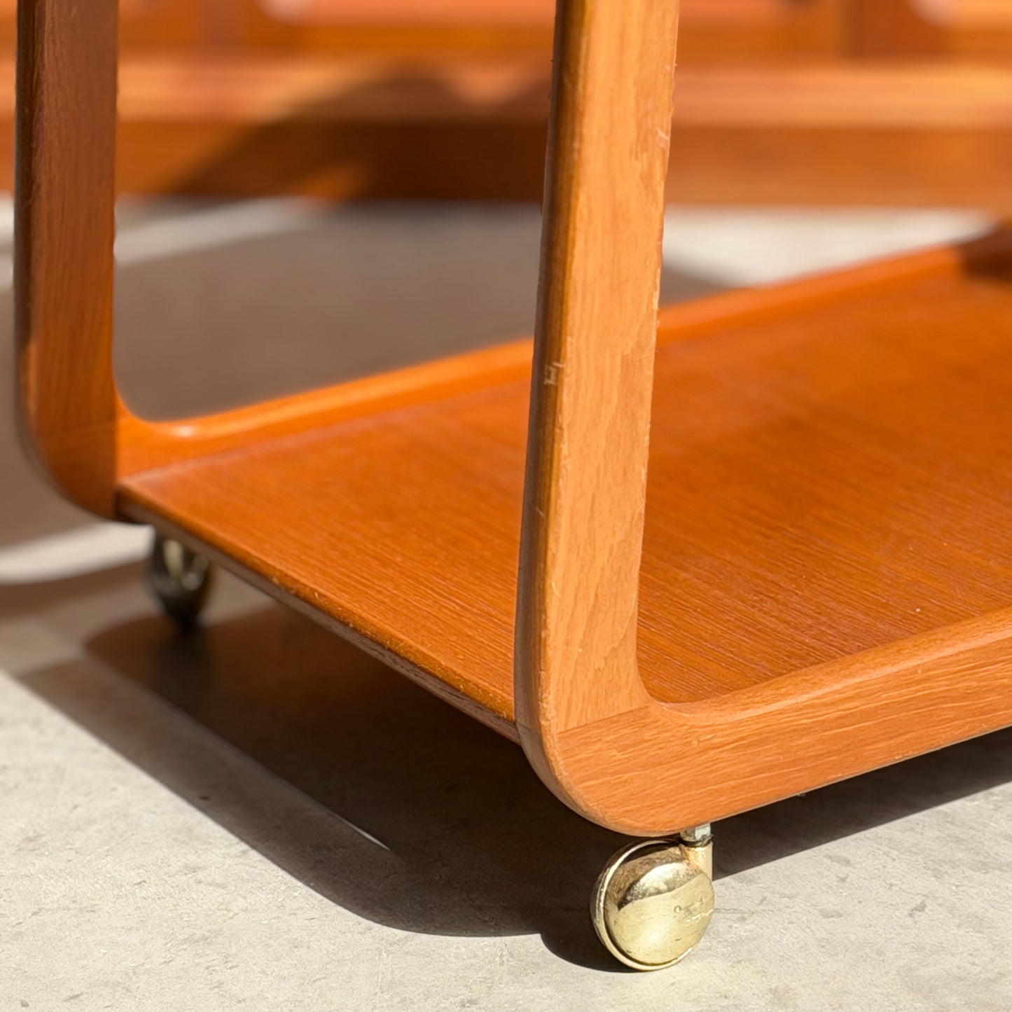 Close-up of the Mid-Century Noblett Drinks Trolley – Removable Tray & Original Castors (Revive), with rounded edges, brass castors, and warm sunlight highlighting its teak grain and smooth finish on a concrete floor.