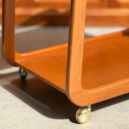 Close-up of the Mid-Century Noblett Drinks Trolley – Removable Tray & Original Castors (Revive), with rounded edges, brass castors, and warm sunlight highlighting its teak grain and smooth finish on a concrete floor.