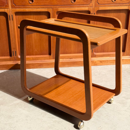 The Mid-Century Noblett Drinks Trolley – Removable Tray & Original Castors (Revive) features a minimalist design with curved edges, two shelves, and four caster wheels. Sunlight casts shadows on the trolley and nearby wooden cabinets.