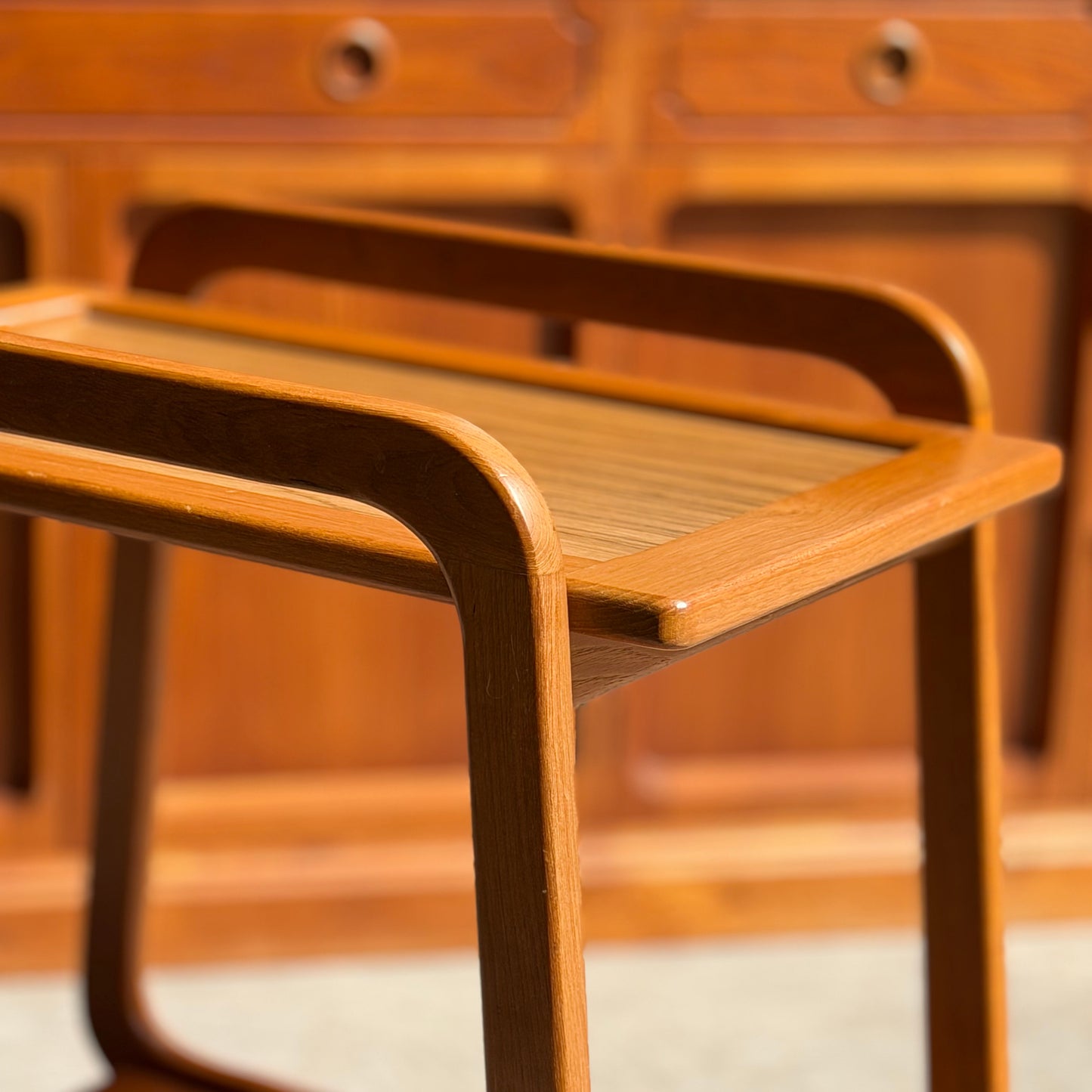 A close-up of the Mid-Century Noblett Drinks Trolley – Removable Tray & Original Castors (Revive), with curved handles and legs, placed by matching cabinets. Sunlight highlights its rich teak grain and classic Noblett craftsmanship.