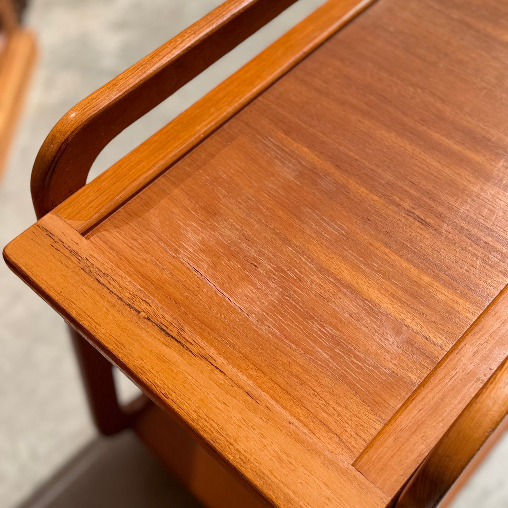 A close-up of a Mid-Century Noblett Drinks Trolley – Removable Tray & Original Castors (Revive) shows its smooth, warm-toned teak finish, rounded edges, visible wood grain, and some light wear and scratches on the surface.