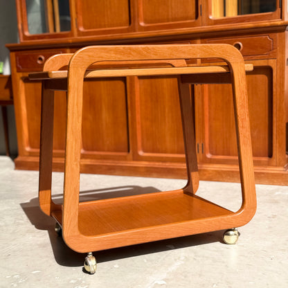 The Mid-Century Noblett Drinks Trolley – Removable Tray & Original Castors (Revive) stands on a sunlit floor before matching wooden cabinetry, showcasing its modern minimalist design with rounded frames, lower shelf, and four caster wheels.