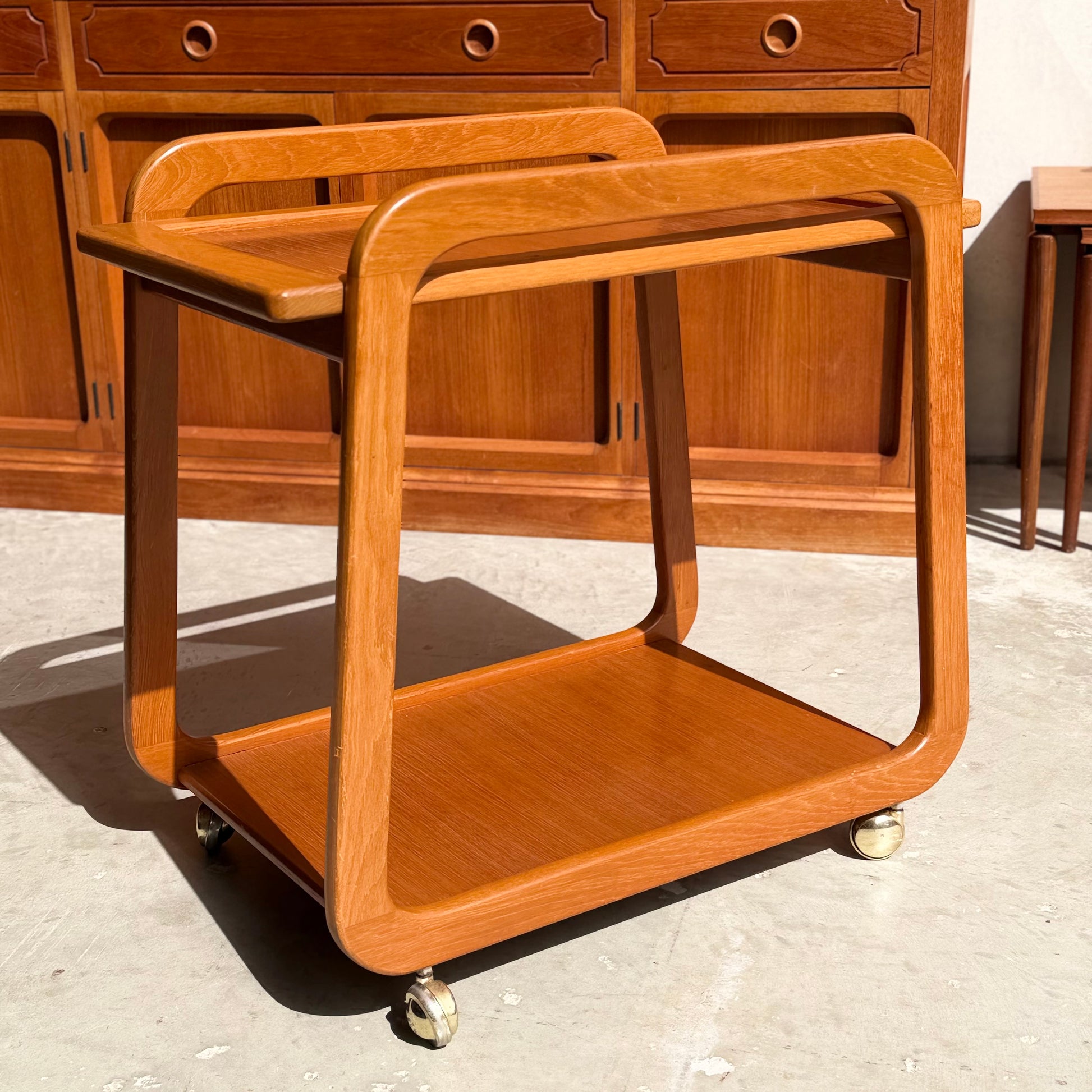 The Mid-Century Noblett Drinks Trolley – Removable Tray & Original Castors (Revive) stands in a sunlit room with matching wooden décor, its classic rounded edges and two-tier design echoing timeless Noblett style.