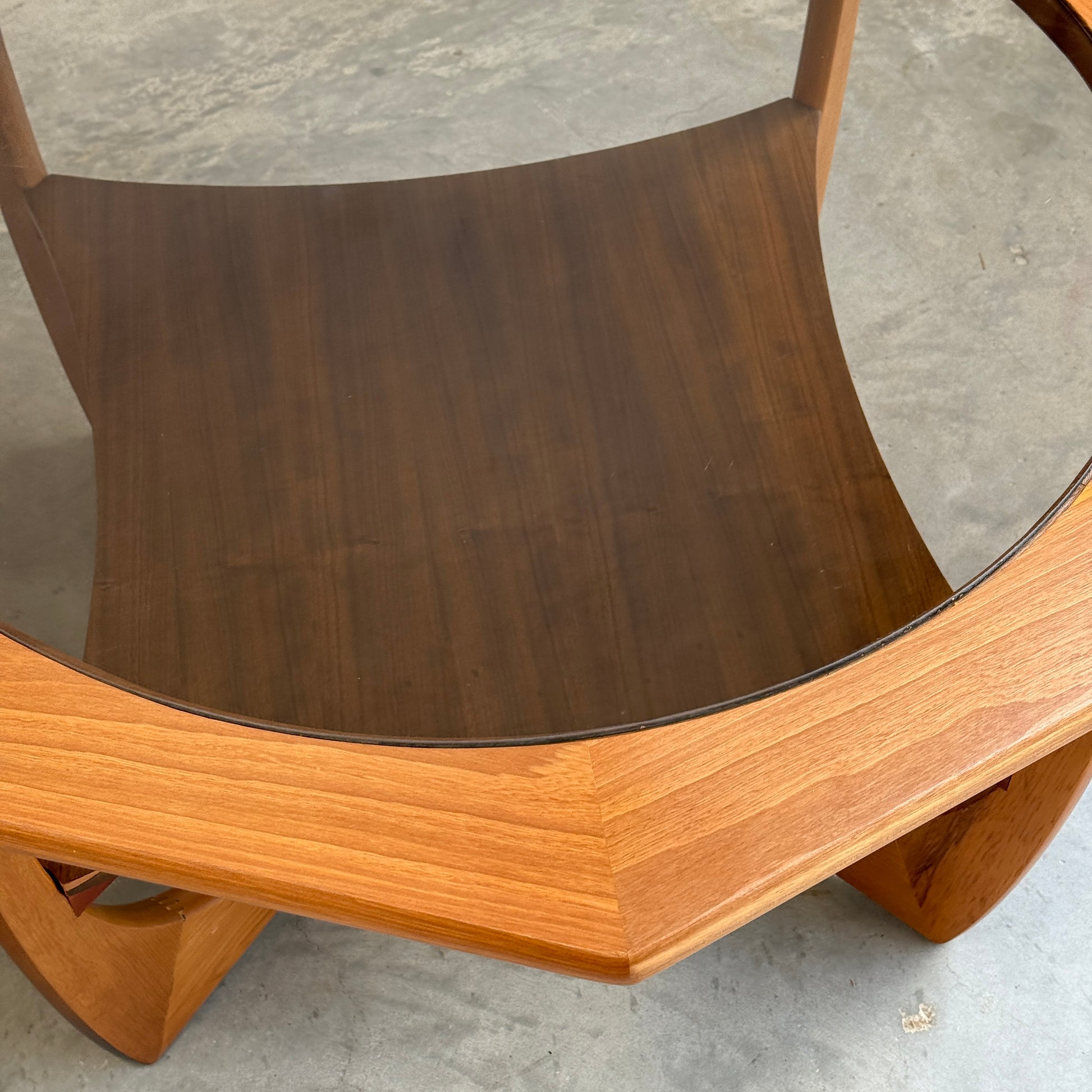 An Octagonal Mid Century Coffee Table by Kalmar Furniture, made of hardwood with a smoked glass top and magazine rack, sits on a concrete floor. Its geometric design and contrasting woods showcase refined Australian craftsmanship.