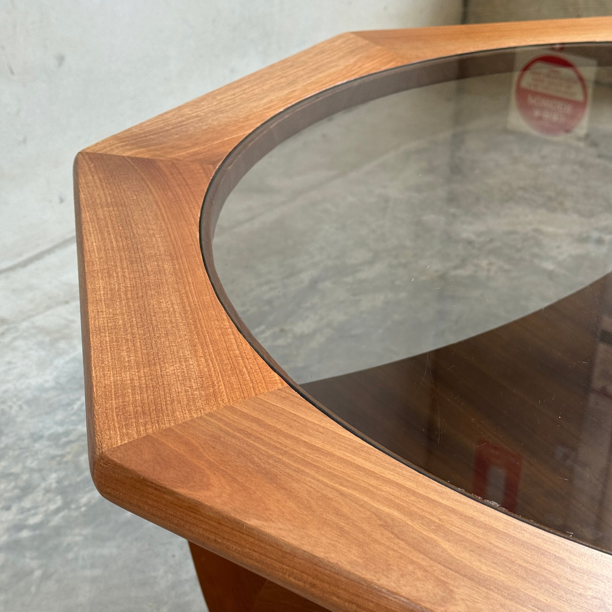 Close-up of the Octagonal Mid Century Coffee Table by Kalmar Furniture, featuring hardwood construction, smoked glass center, and an integrated magazine rack. Its refined joinery and materials evoke classic mid-century style on a concrete floor.
