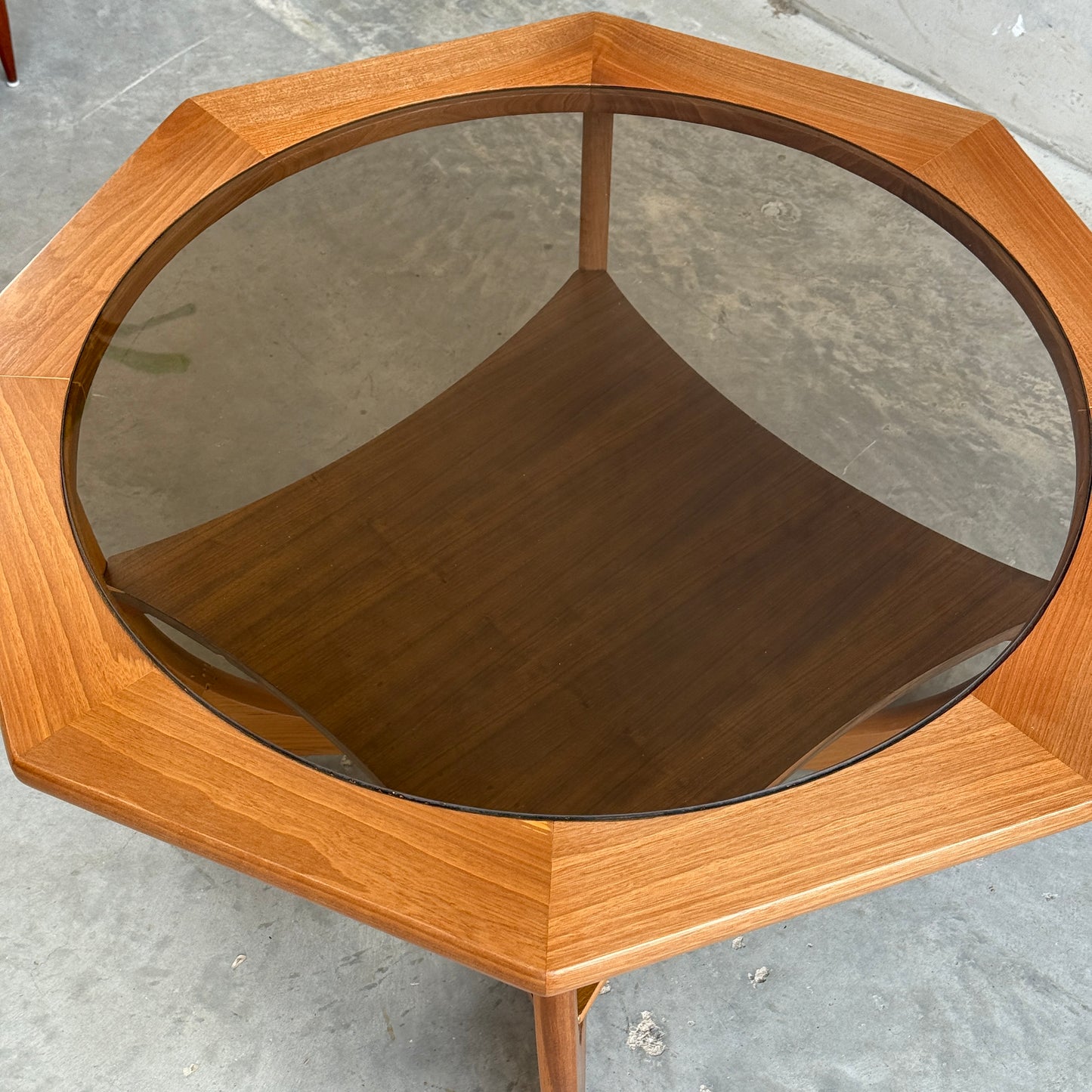 An Octagonal Mid Century Coffee Table by Kalmar Furniture, featuring hardwood construction, a smoked glass top, and a magazine rack, stands on three legs atop a gray concrete floor.