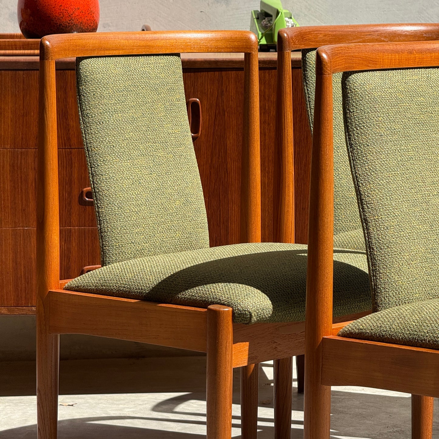 Six fully restored Parker T-Back Number 88 dining chairs in forest green bask in sunlight, while a wooden sideboard with curved handles and a red vase complete the mid-century modern look in the background.