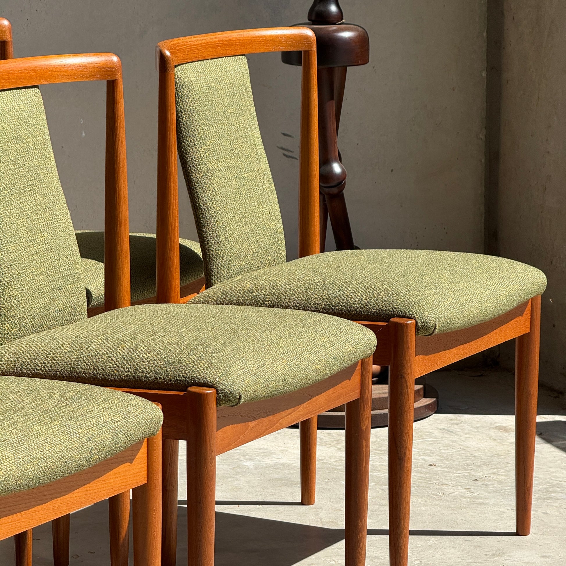 Six fully restored Parker T-Back Number 88 dining chairs in forest green upholstery are bathed in sunlight, creating shadows on a concrete floor. The timeless set from Rediscover stands out against a beige wall.
