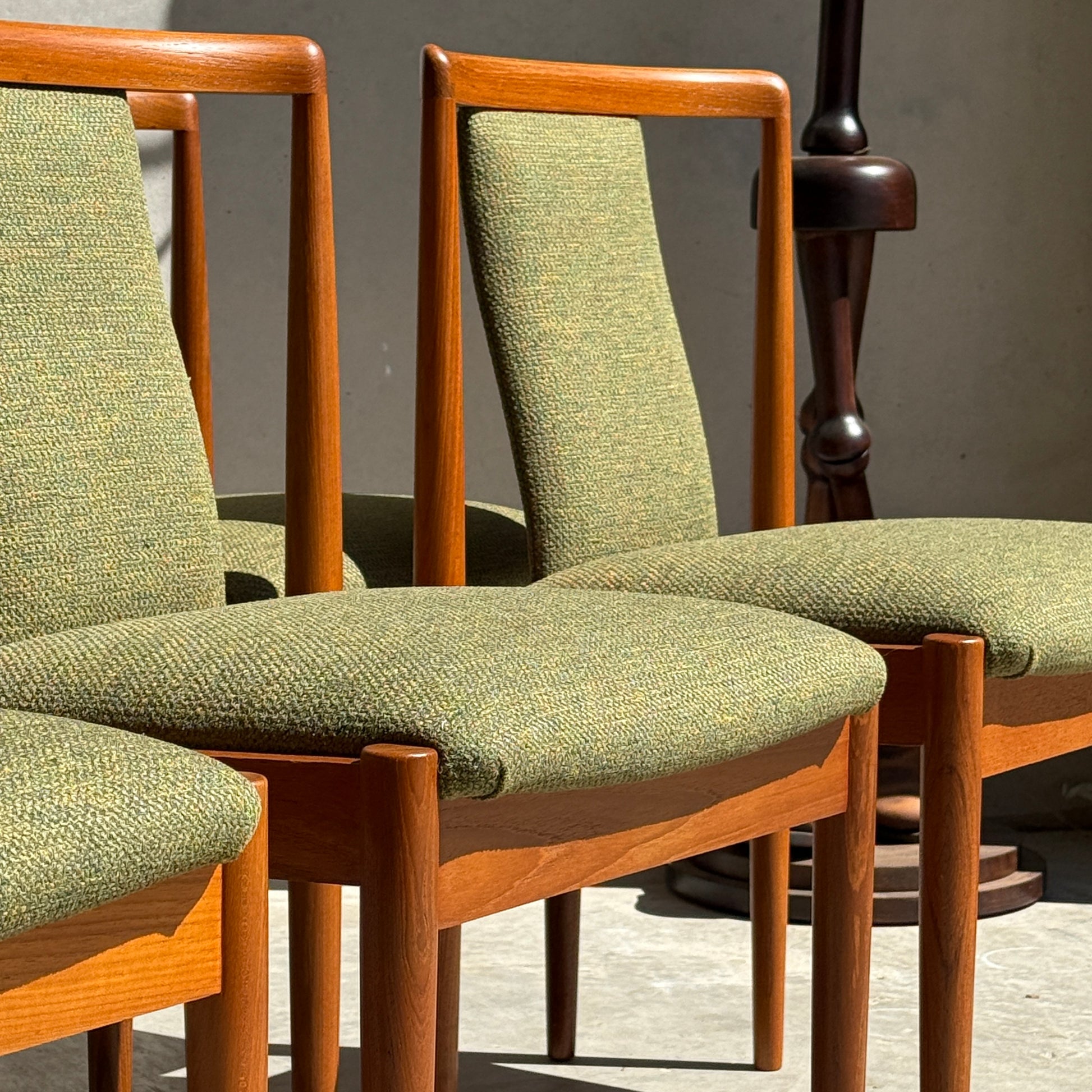 Six fully restored Parker T-Back Number 88 dining chairs in forest green are arranged side by side in sunlight, casting soft shadows. A dark wooden stand appears in the background—timeless elegance from Rediscover Furniture.
