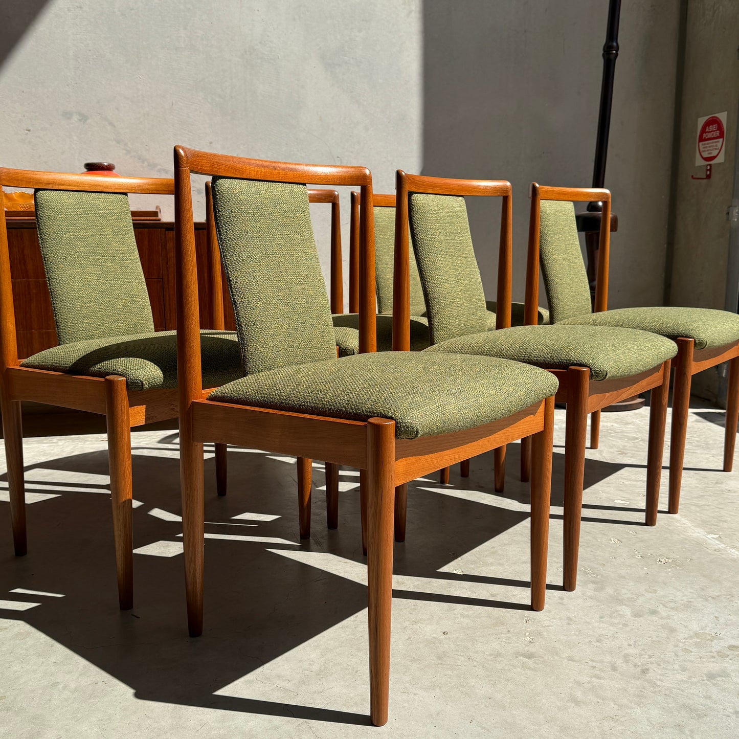 A set of six fully restored Parker T-Back Number 88 dining chairs in forest green upholstery is arranged in a row on a concrete floor, sunlight streaming against a plain, light-colored wall.