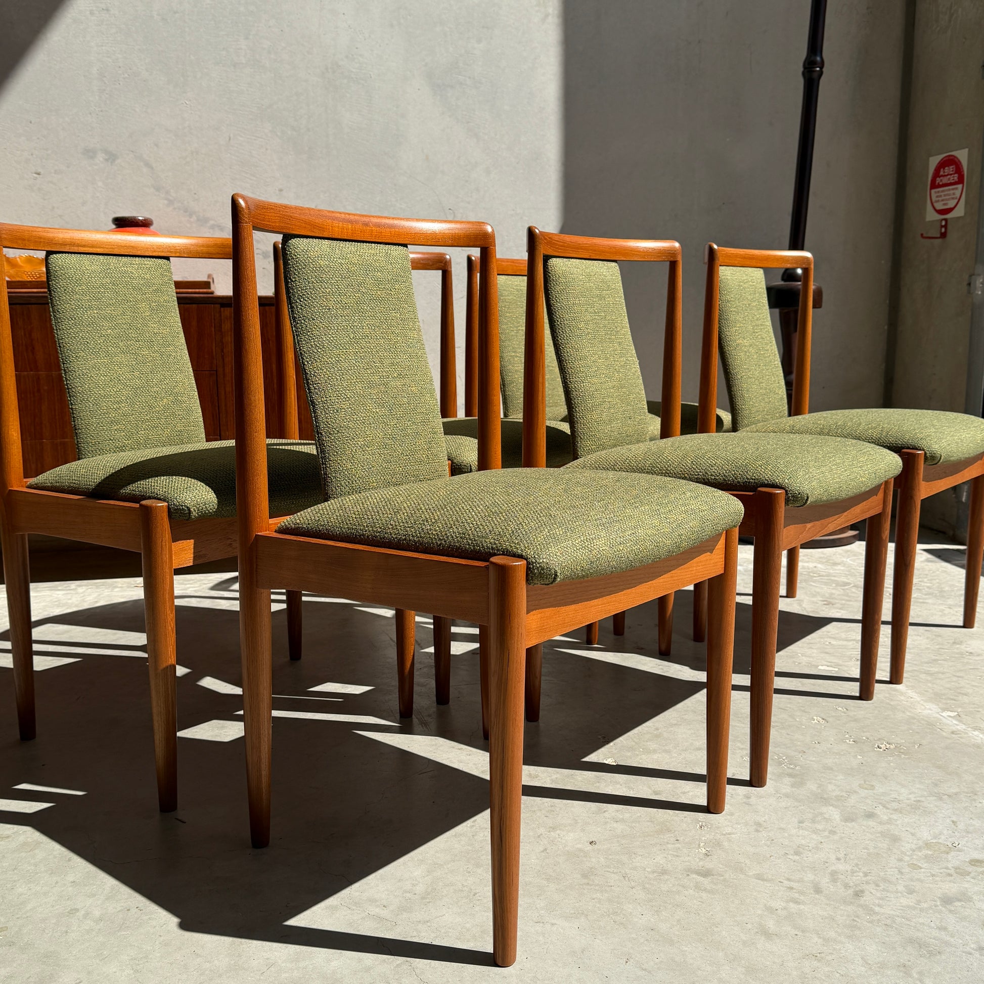 A set of six fully restored Parker T-Back Number 88 dining chairs in forest green upholstery is arranged in a row on a concrete floor, sunlight streaming against a plain, light-colored wall.