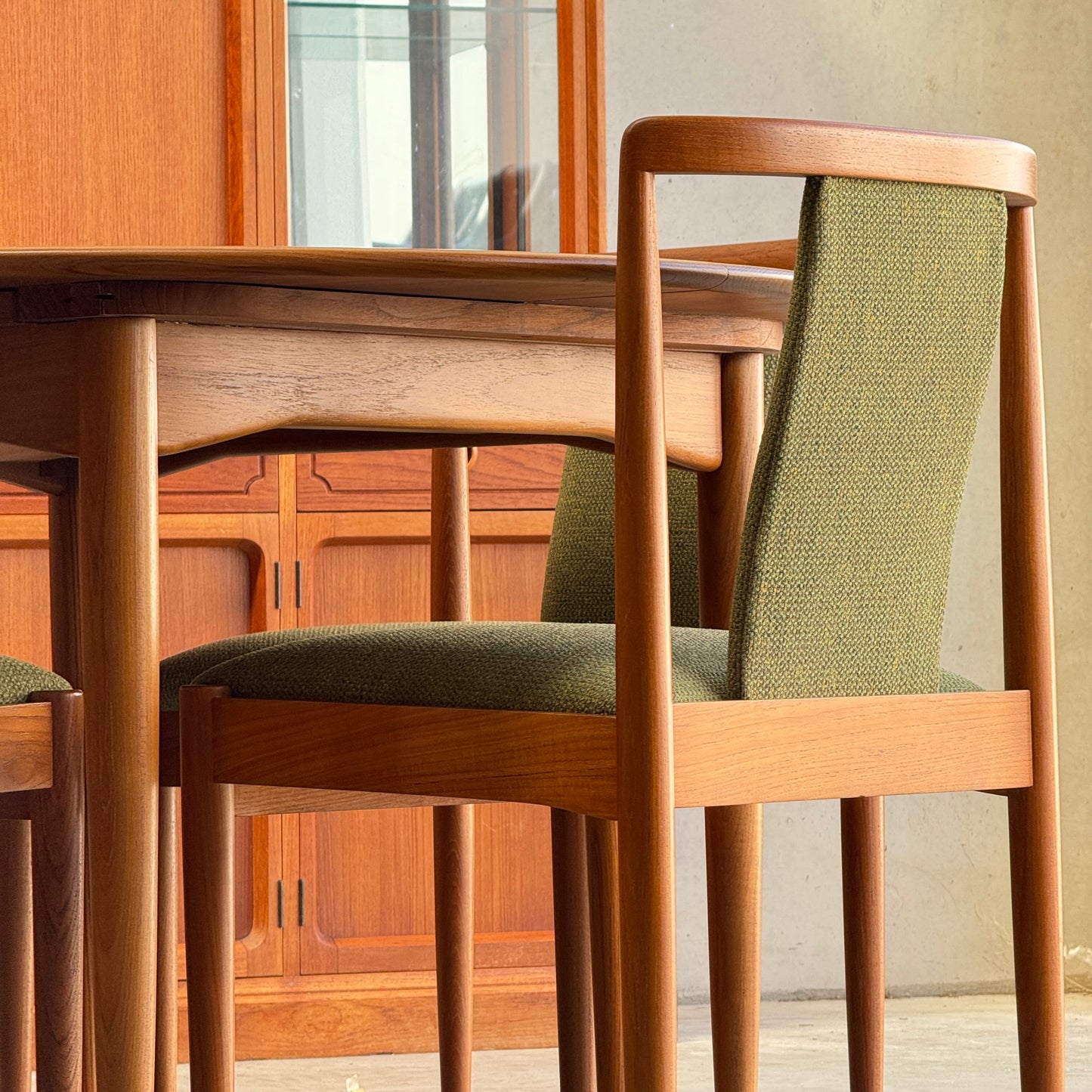Two fully restored Parker T-Back Number 88 dining chairs in forest green upholstery from a set of six (Rediscover) sit by a matching table, with a wooden cabinet featuring glass doors visible against the neutral wall behind them.