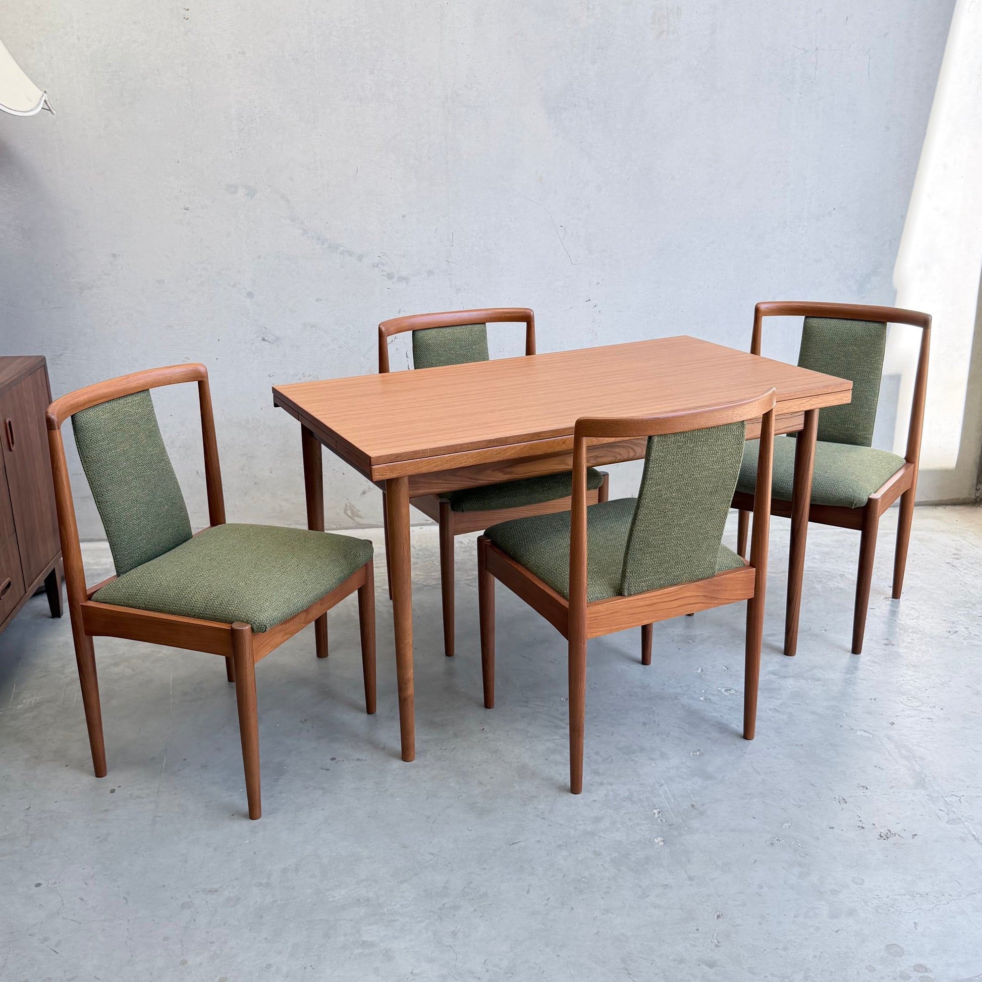 A wooden dining table with a set of Fully Restored Parker T-Back Number 88 Dining Chairs (Set of 4 - Forest Green, Rediscover) sits on a concrete floor against a plain light gray wall.