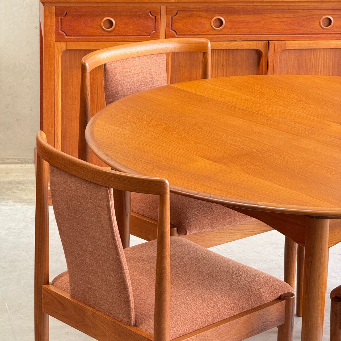 A round wooden dining table is paired with a set of four fully restored Parker T-Back Number 88 Dining Chairs in terracotta, placed before a wooden sideboard with drawers for a simple mid-century modern look.
