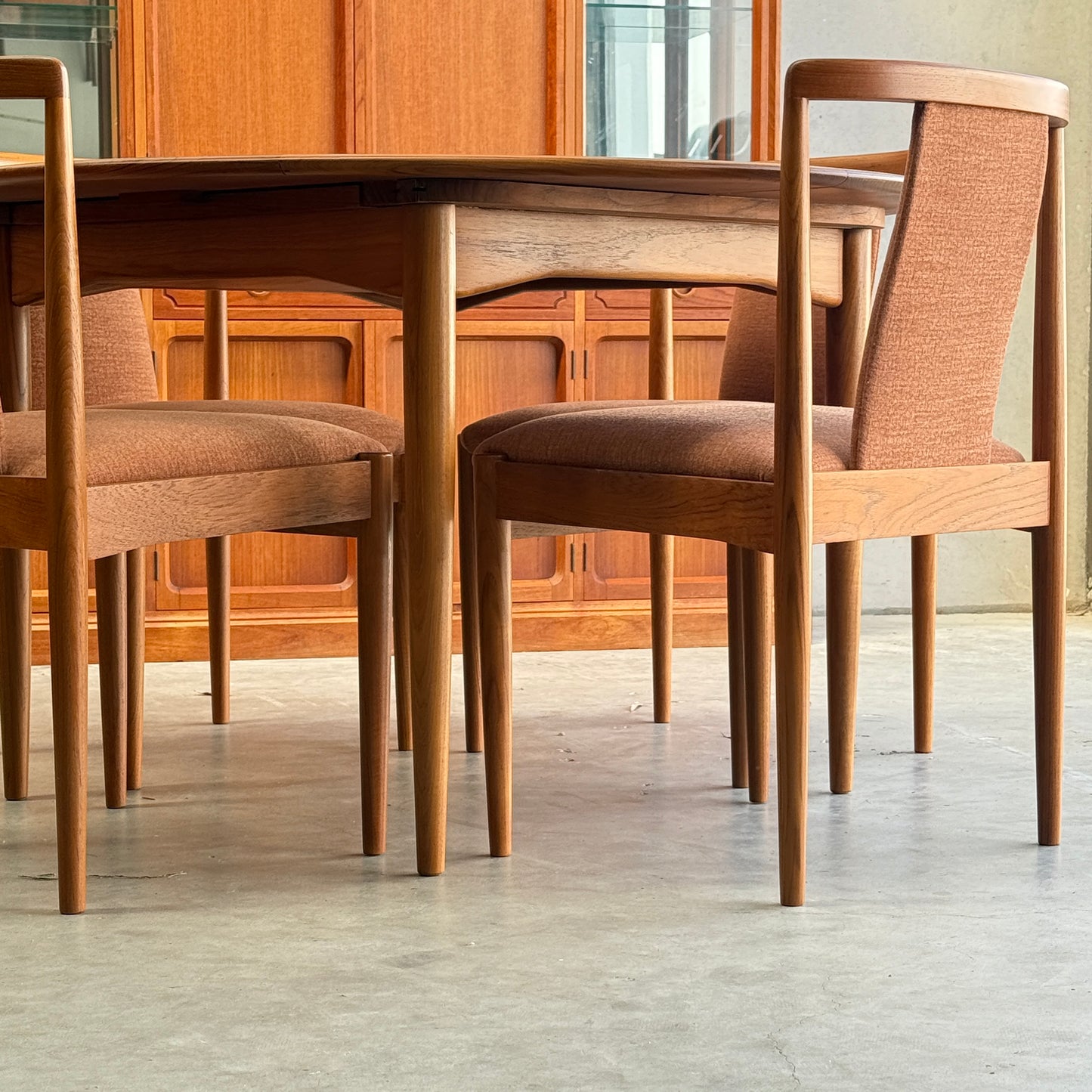Four fully restored Parker T-Back Number 88 dining chairs in terracotta surround a matching wooden dining table on a concrete floor, with a wooden cabinet and glass display case visible in the background.