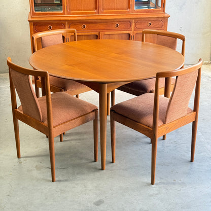 A round wooden dining table with a set of four fully restored Parker T-Back Number 88 Dining Chairs in terracotta upholstery. In the background, a wooden cabinet with drawers and glass doors stands against a concrete wall.