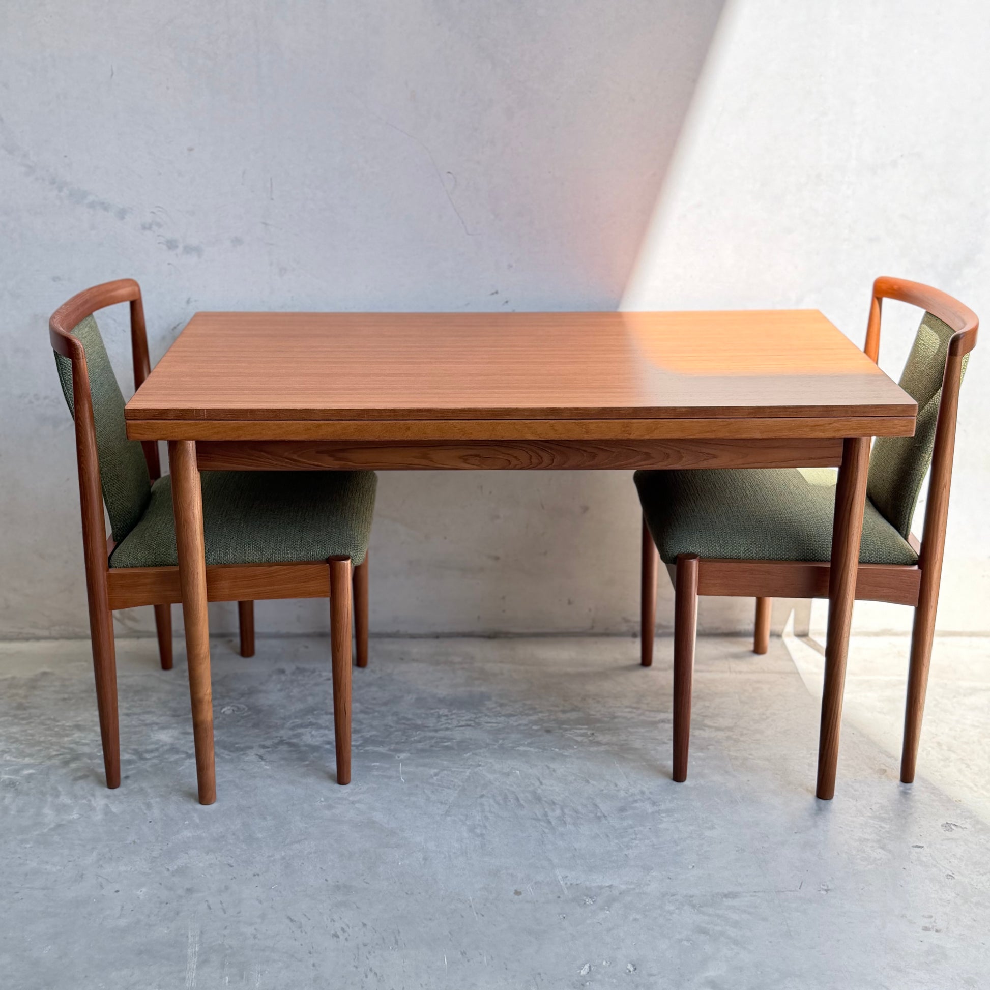 A wooden dining table with a rectangular top and four fully restored Parker T-Back Number 88 Dining Chairs in forest green, set against a light wall and concrete floor for a mid-century modern vibe.