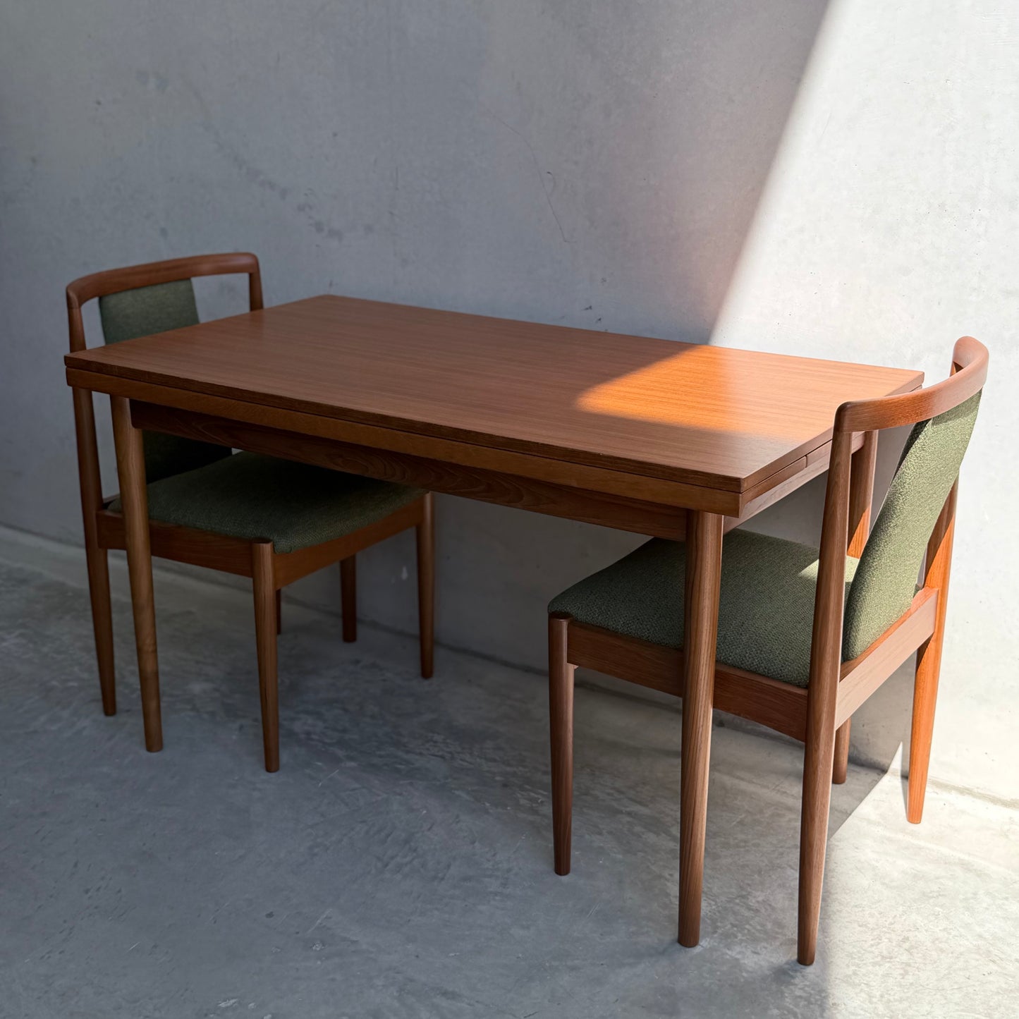 The Fully Restored Parker Dining Suite 5 Piece - Forest Green (Rediscover) features a Scandinavian-inspired wooden table and chairs with green upholstery, set against a light gray textured wall in natural sunlight.