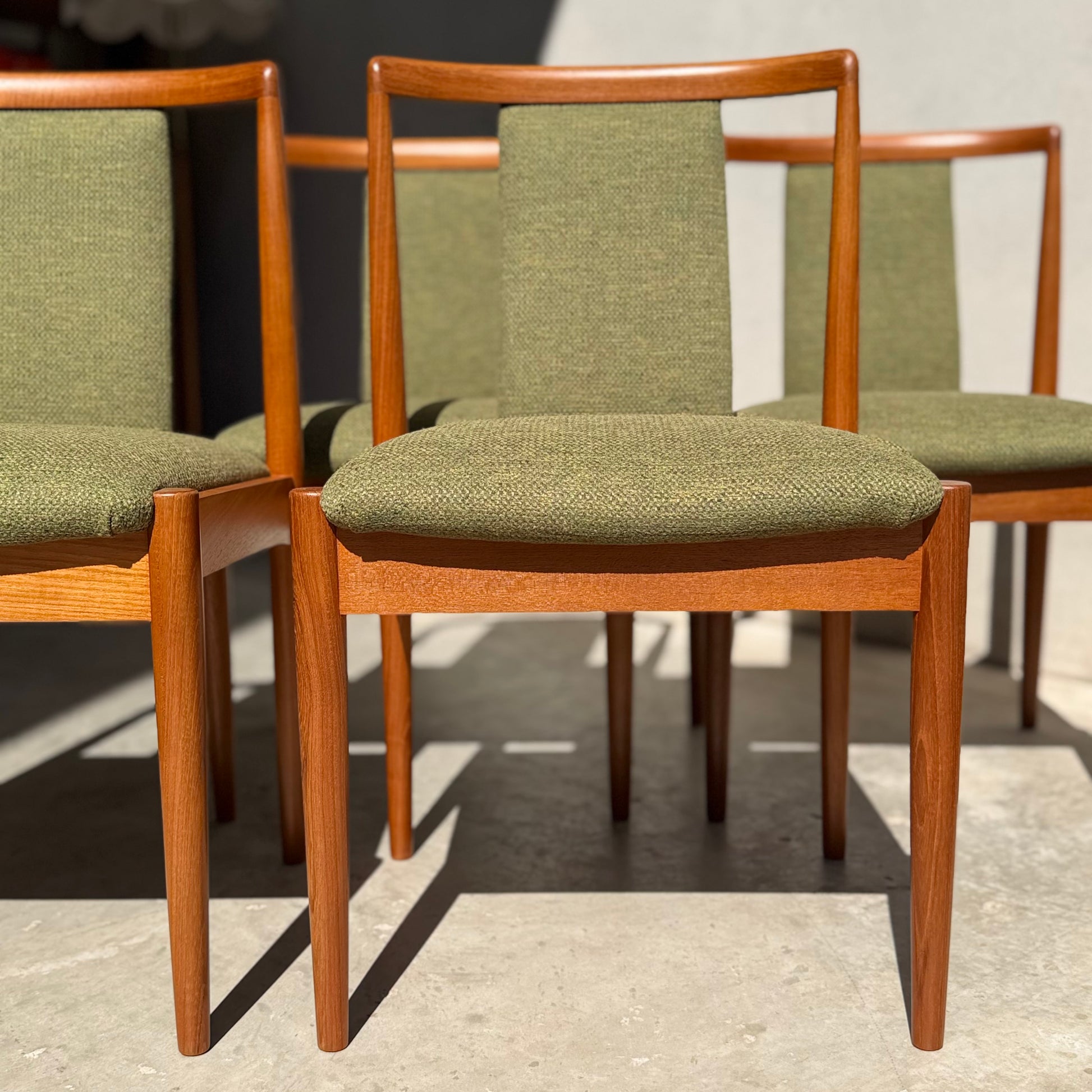 A set of four fully restored Parker T-Back Number 88 dining chairs in forest green from Rediscover are closely arranged on a concrete floor, sunlight casting elegant shadows behind their mid-century modern teak frames.