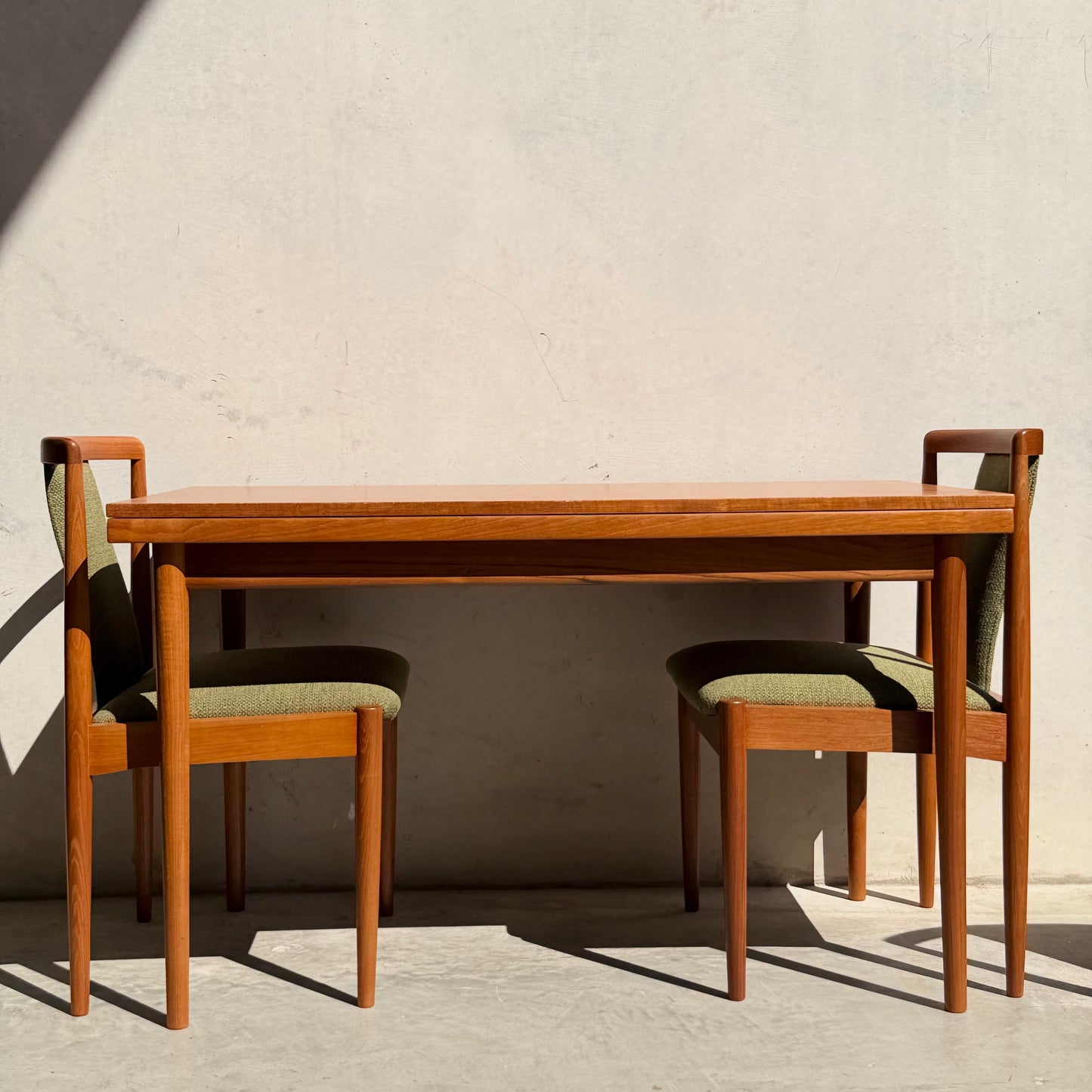 A fully restored Parker Dining Suite, 5-piece set in forest green (Rediscover), features a Scandinavian-inspired wooden table and chairs with green upholstery, set against a plain, light wall with a shadow on the left.