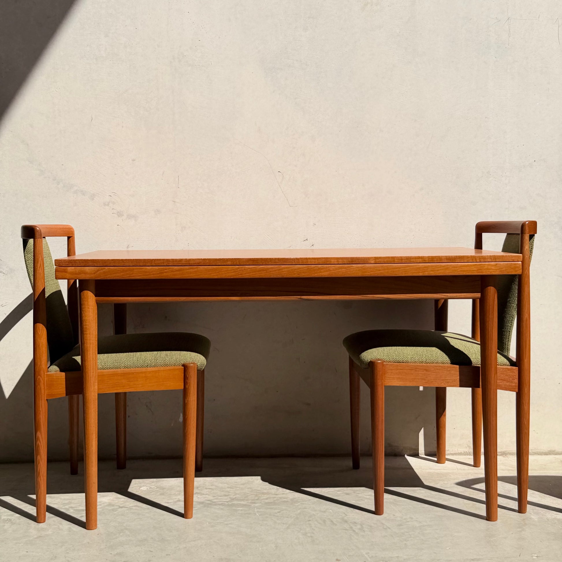 A fully restored Parker Dining Suite, 5-piece set in forest green (Rediscover), features a Scandinavian-inspired wooden table and chairs with green upholstery, set against a plain, light wall with a shadow on the left.