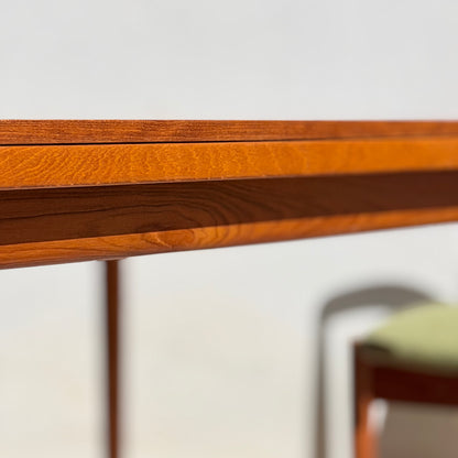 Close-up of the edge of a Fully Restored Parker Dining Suite table with a smooth, reddish-brown finish. In the background, a softly blurred chair with a forest green cushion from this 5-piece set is visible in the well-lit room.