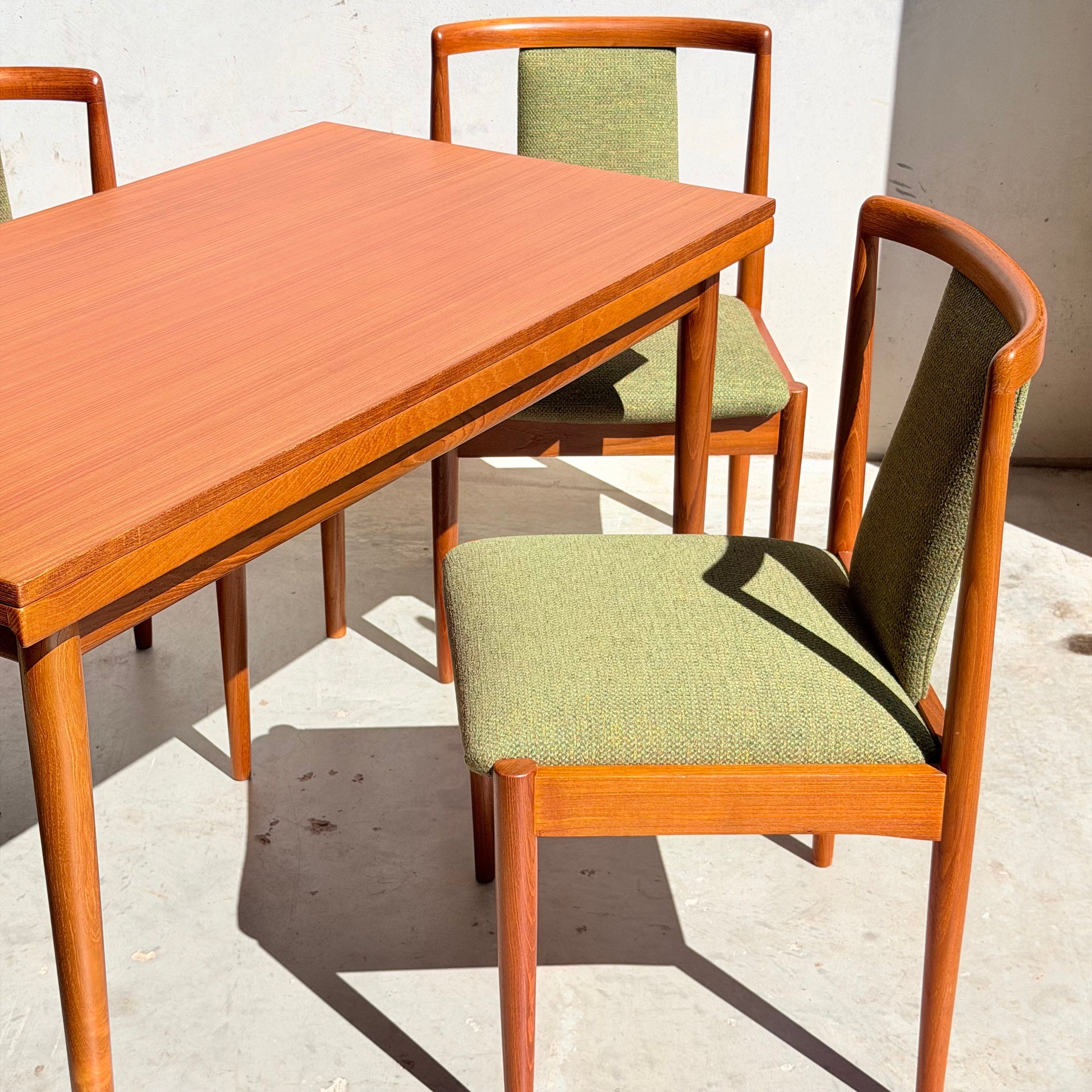A Fully Restored Parker Dining Suite 5 Piece – Forest Green (Rediscover) is arranged on a sunlit concrete floor, featuring sleek mid-century modern chairs with green upholstery around a matching rectangular table.