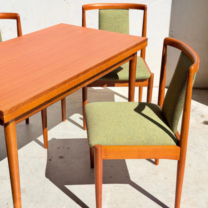 A Fully Restored Parker Dining Suite 5 Piece – Forest Green (Rediscover) is arranged on a sunlit concrete floor, featuring sleek mid-century modern chairs with green upholstery around a matching rectangular table.