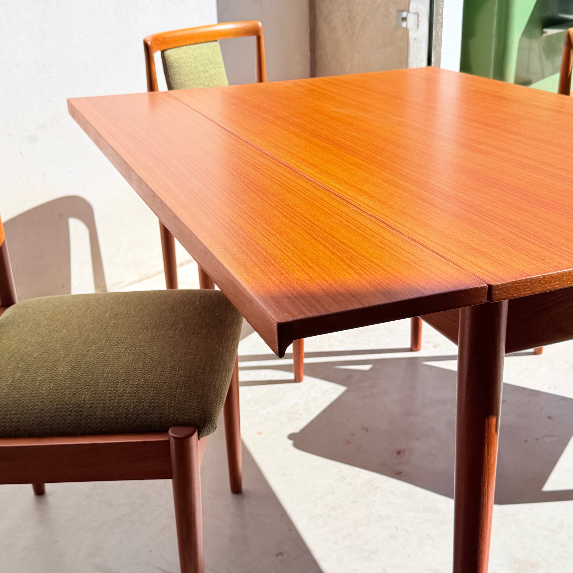 A close-up of the Fully Restored Parker Dining Suite 5 Piece - Forest Green, featuring a wooden table with a rich finish and matching green upholstered chairs, bathed in sunlight—an inviting mid-century modern set.