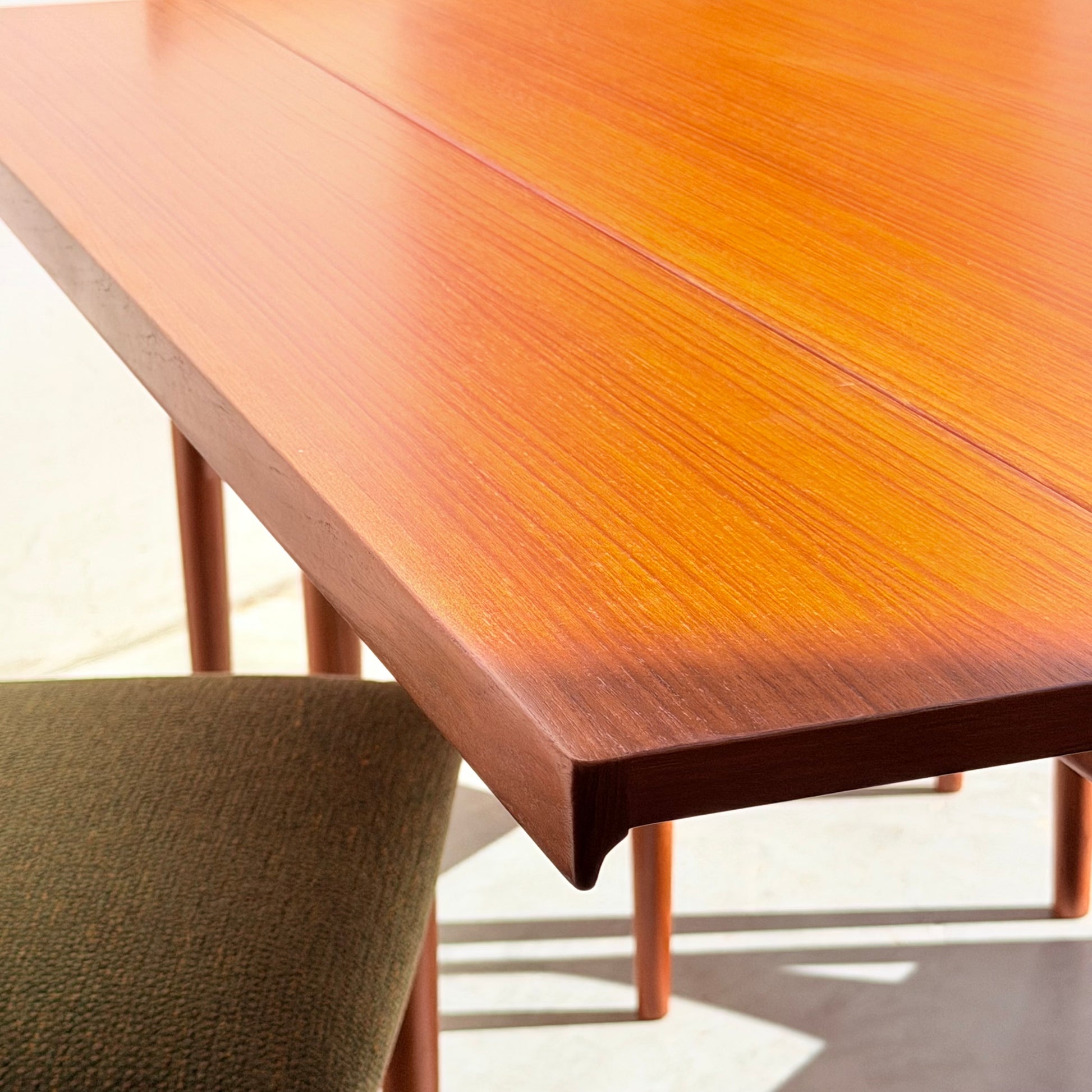 A close-up of the Fully Restored Parker Dining Suite 5 Piece - Forest Green shows its warm wood finish and clean lines beside a green upholstered chair, with sunlight highlighting the Scandinavian-inspired grain.