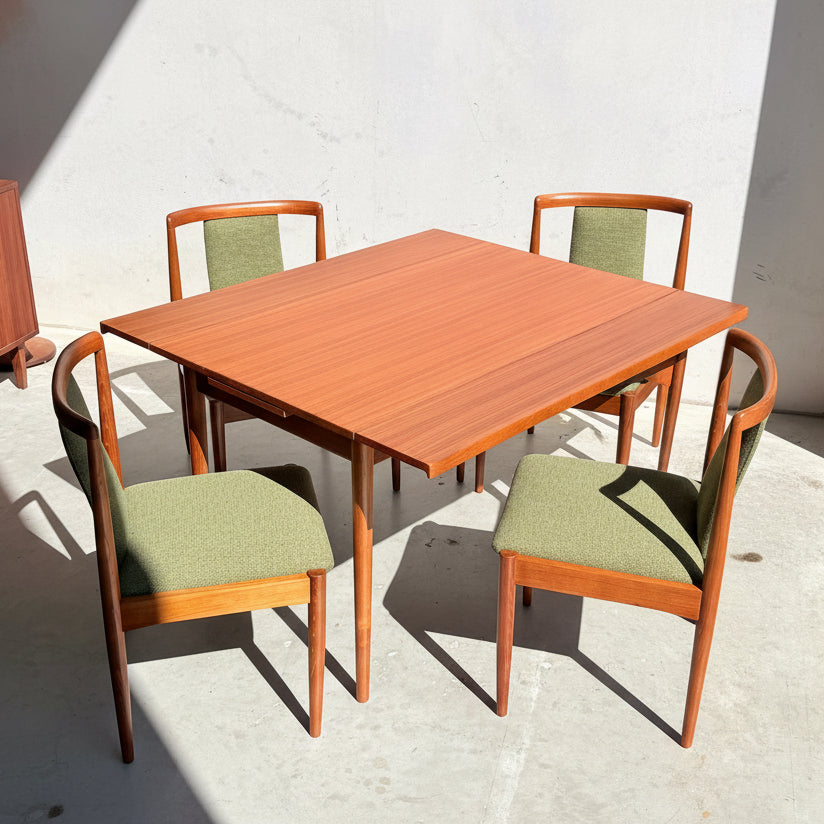 Featuring four forest green upholstered chairs, the Fully Restored Parker Dining Suite 5 Piece (Rediscover) showcases a square wooden table with a reddish-brown finish, set on a light concrete floor dappled with sunlight.