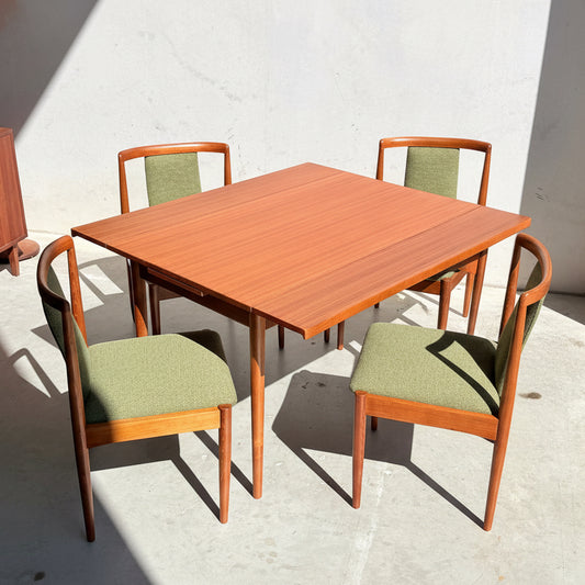 Featuring four forest green upholstered chairs, the Fully Restored Parker Dining Suite 5 Piece (Rediscover) showcases a square wooden table with a reddish-brown finish, set on a light concrete floor dappled with sunlight.