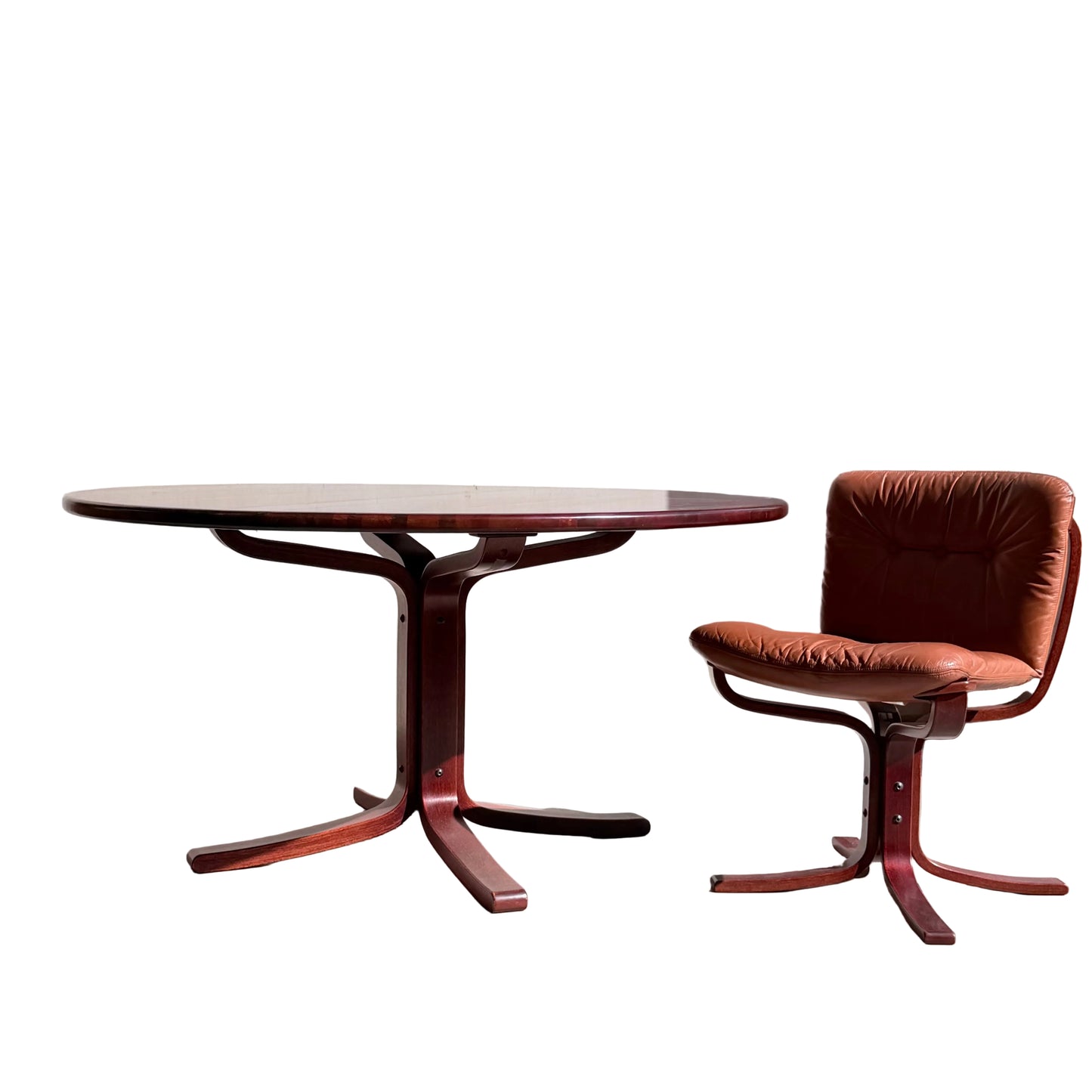 A Joe Rufenacht Designer Round Dining Table – Rare Mid-Century Gem in Solid Wood stands beside a brown cushioned chair with a matching wooden base, set against a plain light wall in natural sunlight.