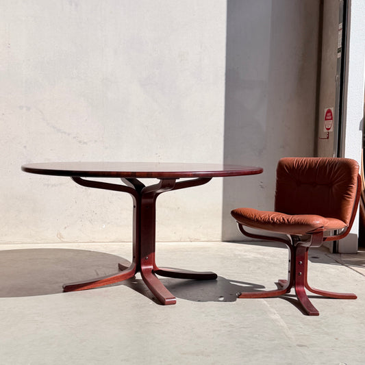 A Joe Rufenacht Designer Round Dining Table – Rare Mid-Century Gem in Solid Wood stands beside a brown cushioned chair with a matching wooden base, set against a plain light wall in natural sunlight.