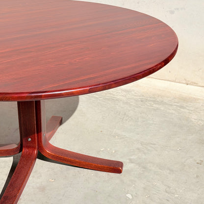A rare Joe Rufenacht Designer Round Dining Table—mid-century solid wood with a polished reddish-brown finish and three-legged base—stands on a light gray concrete floor against a plain, light-colored wall.