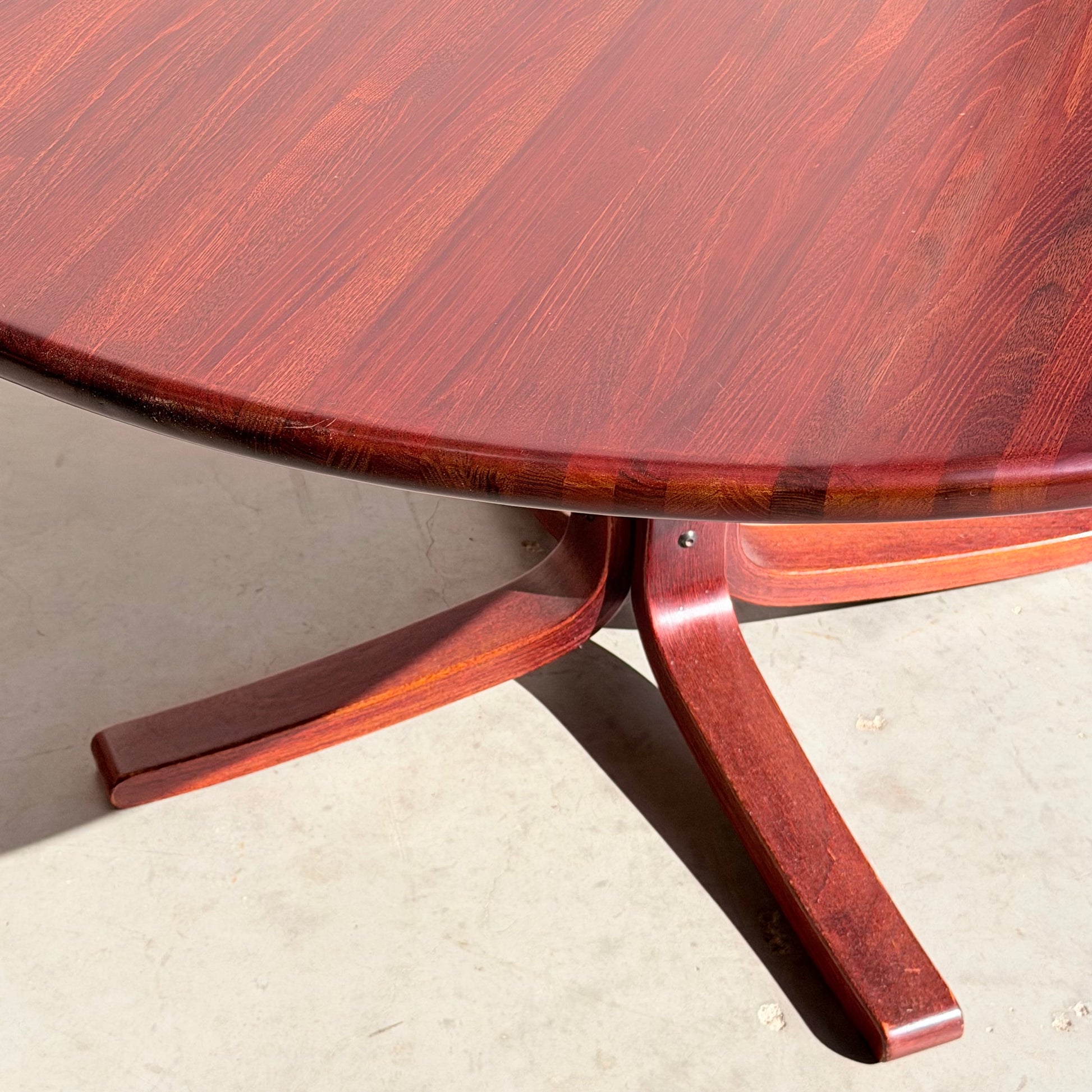 A close-up of the Joe Rufenacht Designer Round Dining Table – Rare Mid-Century Gem in Solid Wood, highlighting its reddish-brown finish and intersecting legs on a light-colored floor.