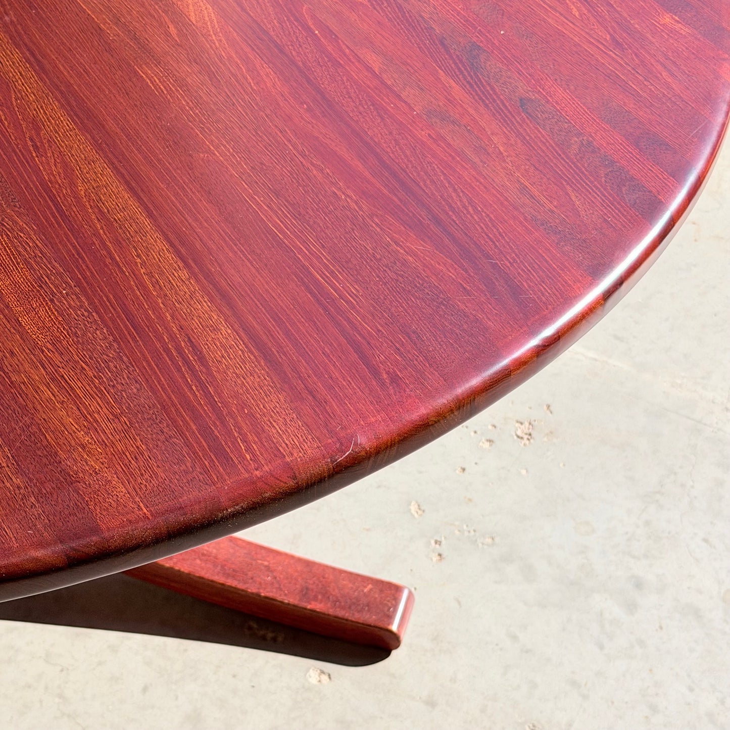 A close-up of the Joe Rufenacht Designer Round Dining Table – Rare Mid-Century Gem in Solid Wood (Refine) showcases its elegant wood grain and part of its leg on a light concrete floor.