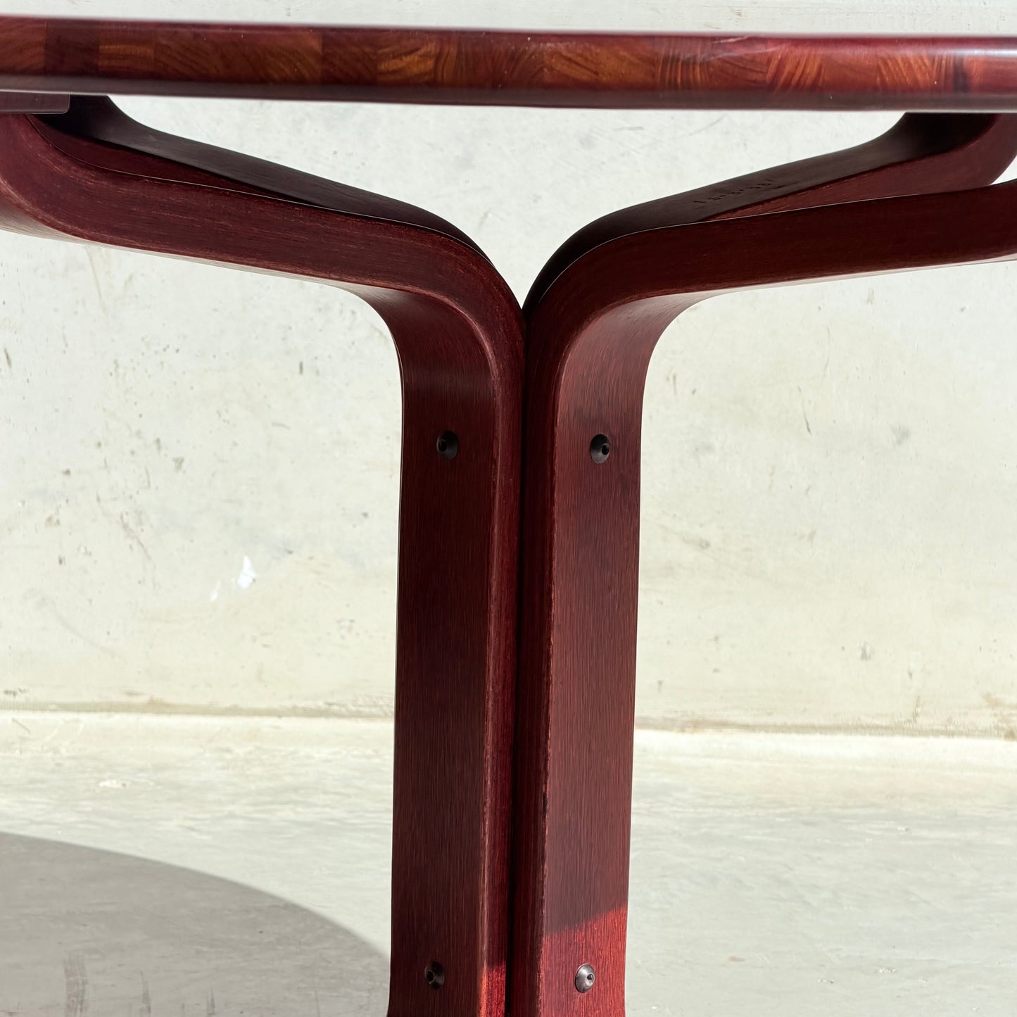 Close-up of the solid wood base and curved, dark reddish legs of the Joe Rufenacht Designer Round Dining Table – Rare Mid-Century Gem in Solid Wood (Refine), joined with screws against a light-colored concrete background.