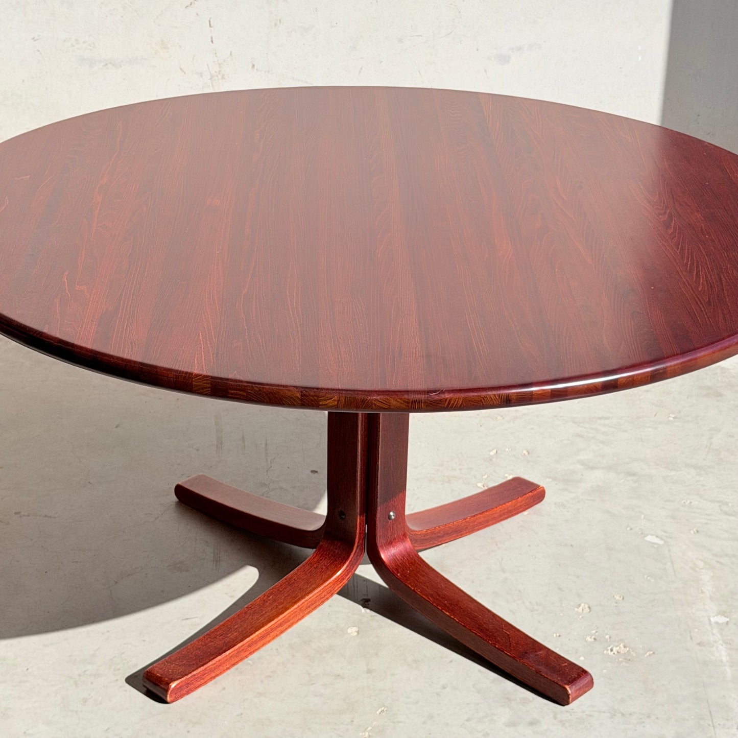The Joe Rufenacht Designer Round Dining Table, a rare mid-century gem in solid wood, features a reddish-brown finish, smooth surface, and sturdy four-pronged base. It stands on a light floor bathed in bright sunlight.
