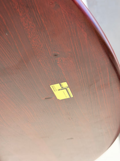 A close-up of the Joe Rufenacht Designer Round Dining Table—Rare Mid-Century Gem in Solid Wood (Refine)—shows its rich wood grain and scratches, with a yellow rectangular sticker featuring a barcode and text attached to the surface.