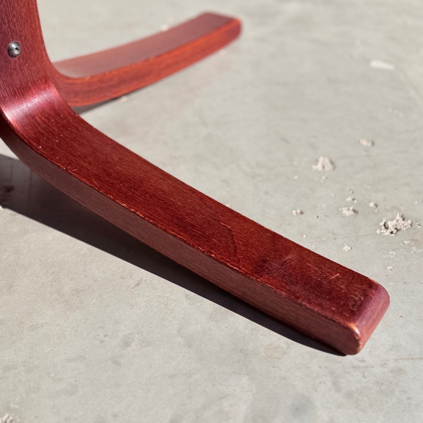 Close-up of a curved, reddish-brown solid wood leg from the Joe Rufenacht Designer Round Dining Table—rare mid-century gem—resting on a light gray, textured concrete floor with small debris scattered nearby.