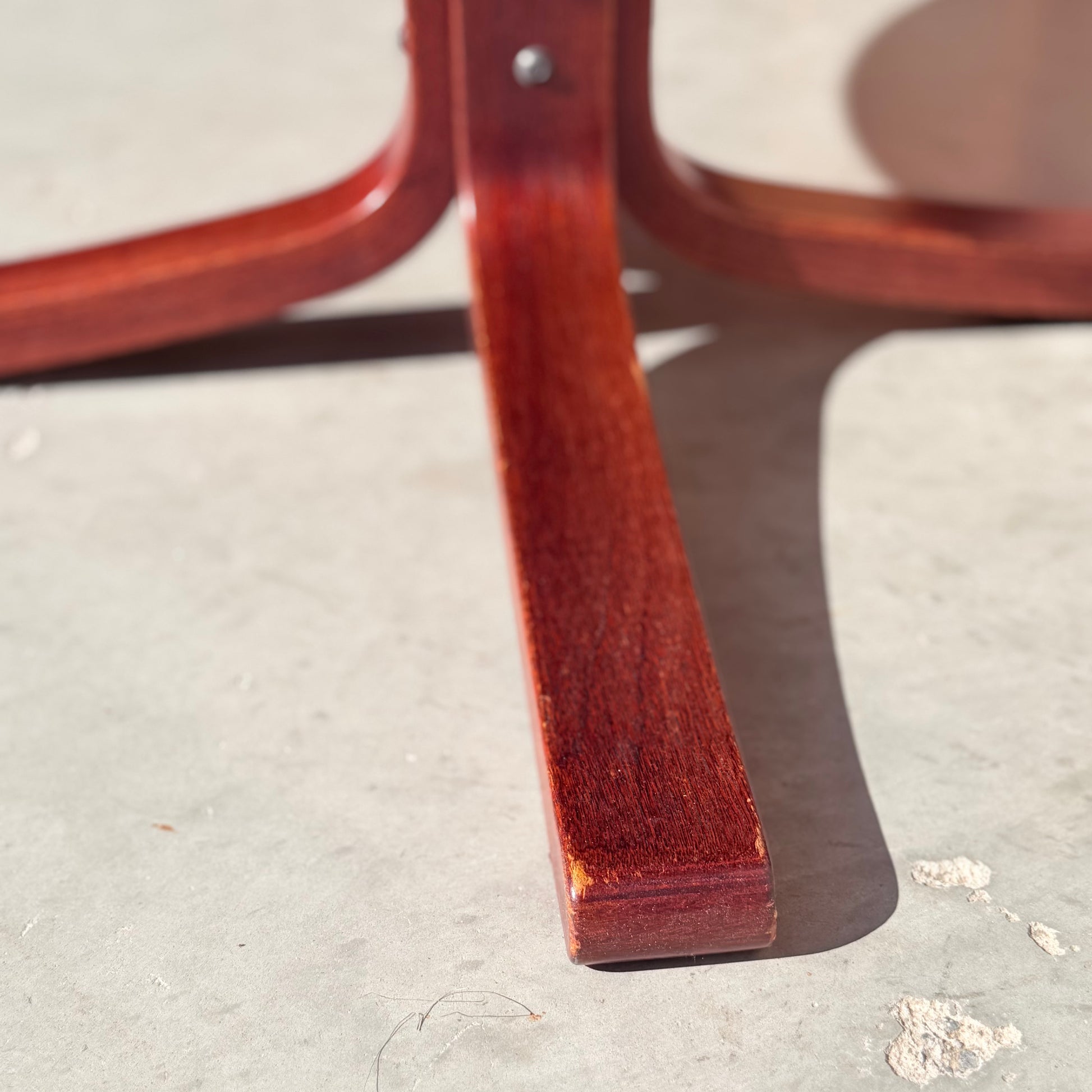 Close-up of the solid wood base of a Joe Rufenacht Designer Round Dining Table – Rare Mid-Century Gem (Refine), featuring a reddish-brown finish on a light gray concrete floor in bright sunlight.